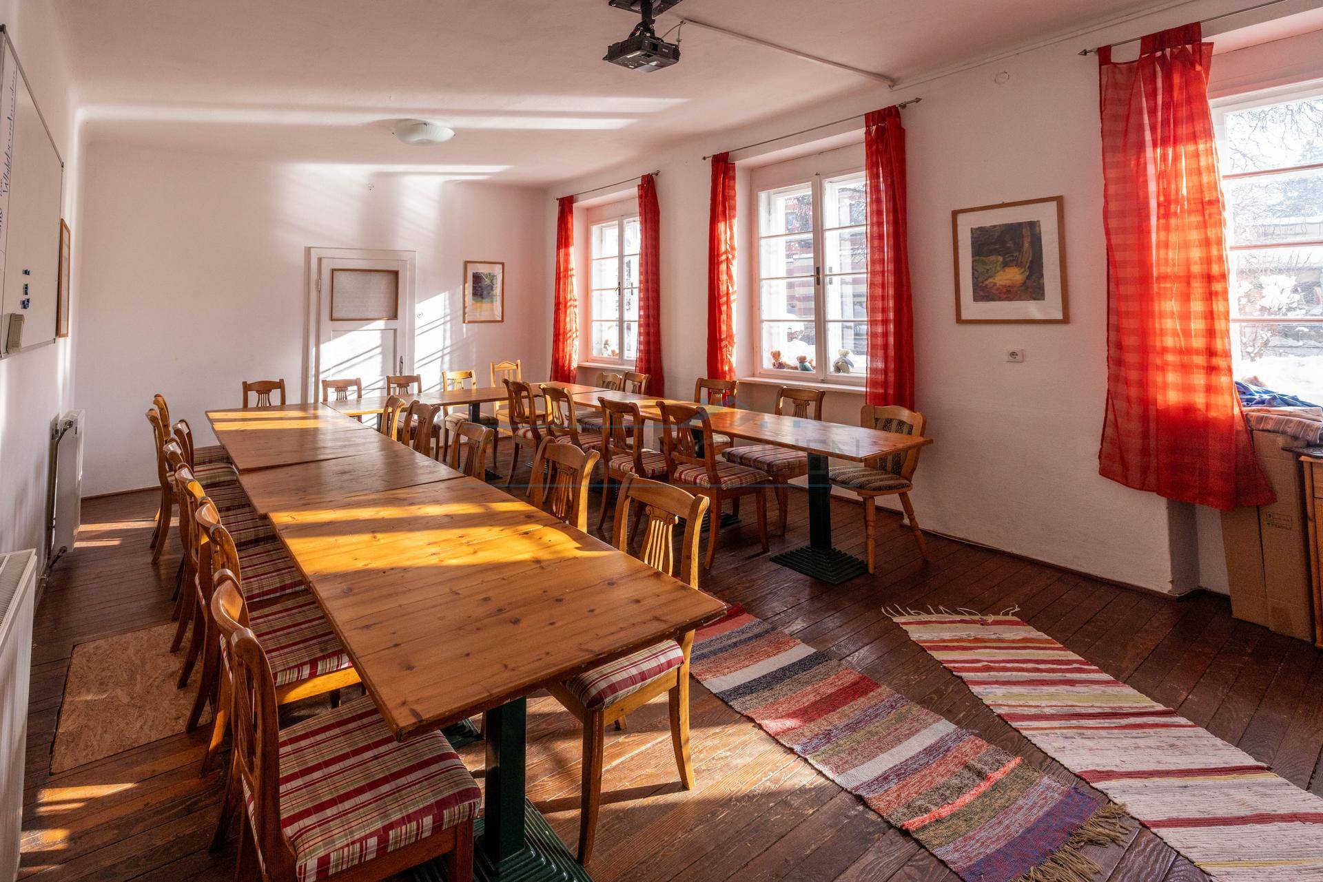 Großer Gemeinschaftsraum mit mehreren Holztischen, Stühlen und roten Vorhängen, ideal für Versammlungen.