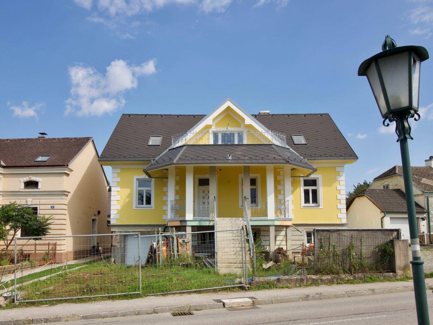 Vorderansicht des gelben Hauses mit Bauzaun und Laterne im sonnigen Tageslicht.