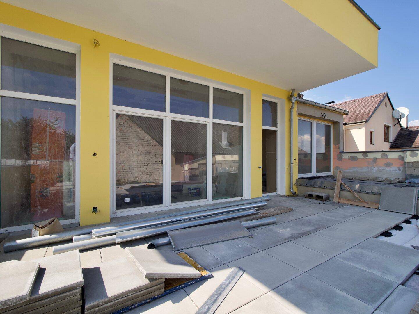 Baustelle einer Terrasse mit großen Fensterfronten und Blick auf Nachbargebäude.
