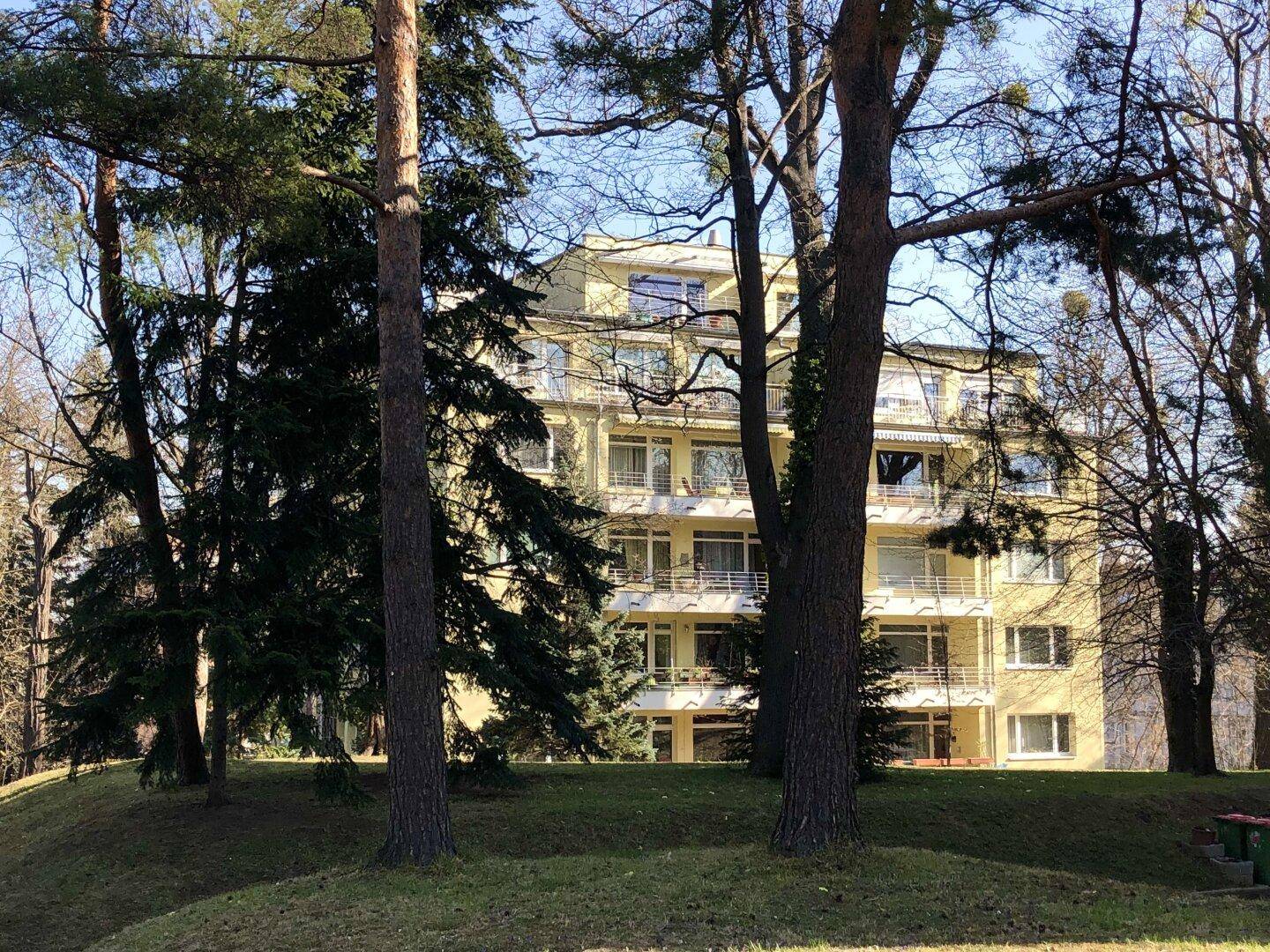 Weitläufiges gelbes Gebäude mit Balkonen, umgeben von hohen Bäumen und grüner Landschaft.