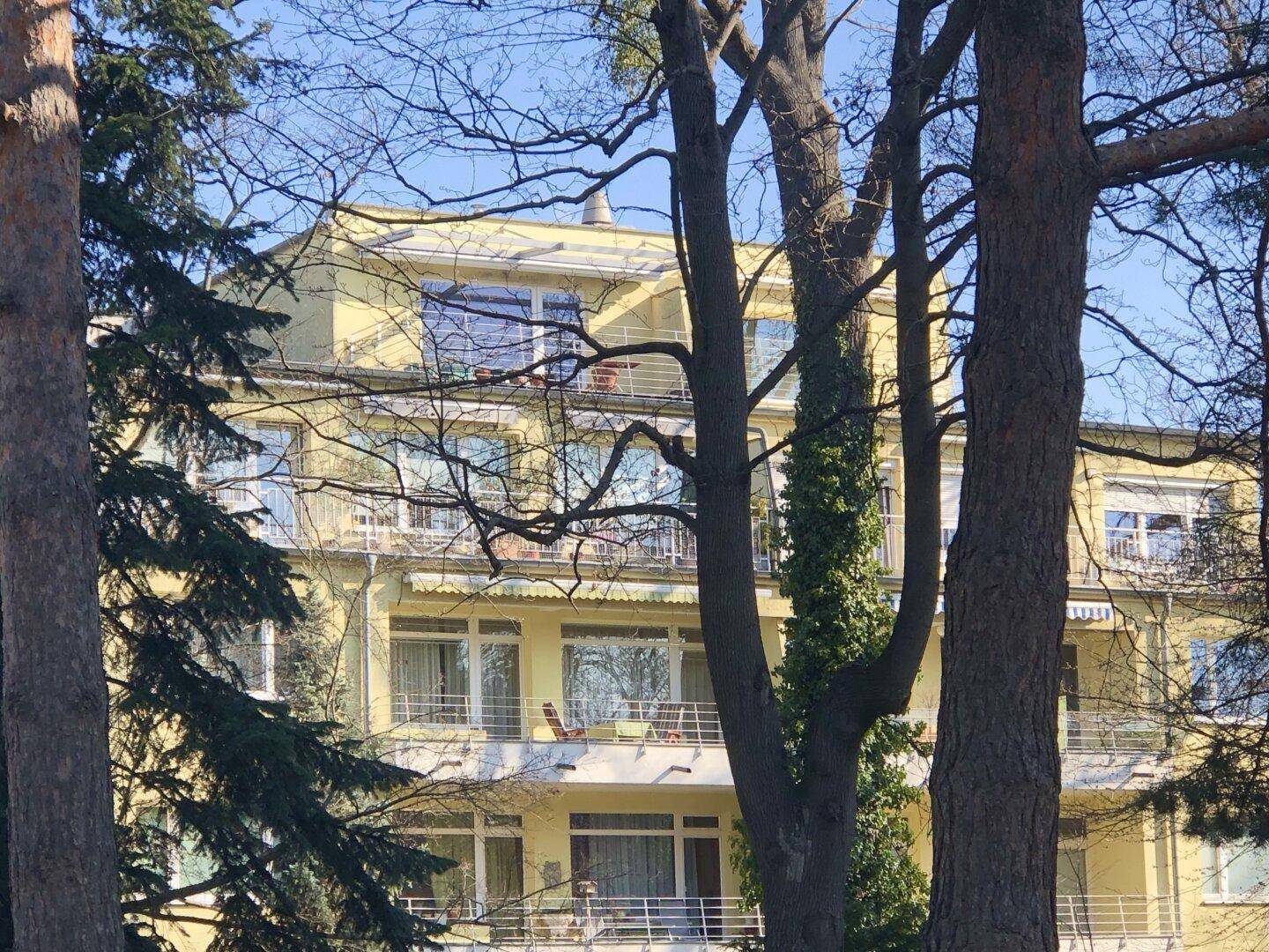 Oberer Teil eines gelben Gebäudes mit mehreren Balkonen, sichtbar durch Baumkronen.