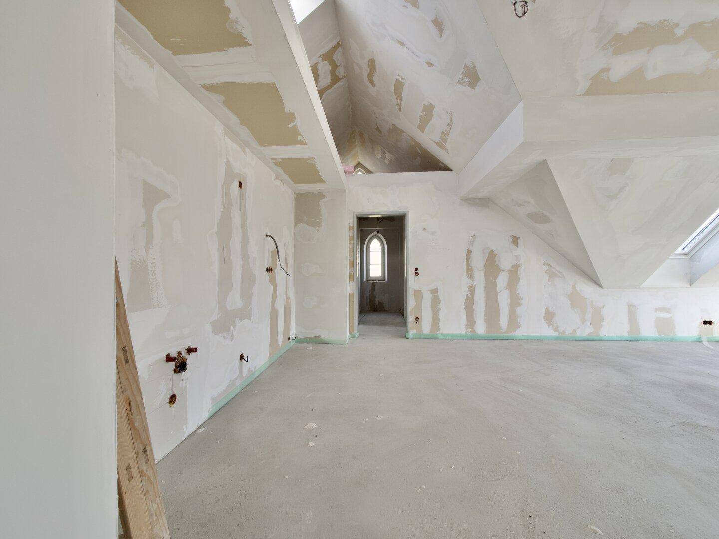Dachboden im Rohzustand mit schrägen Wänden und einem kleinen Bogenfenster.
