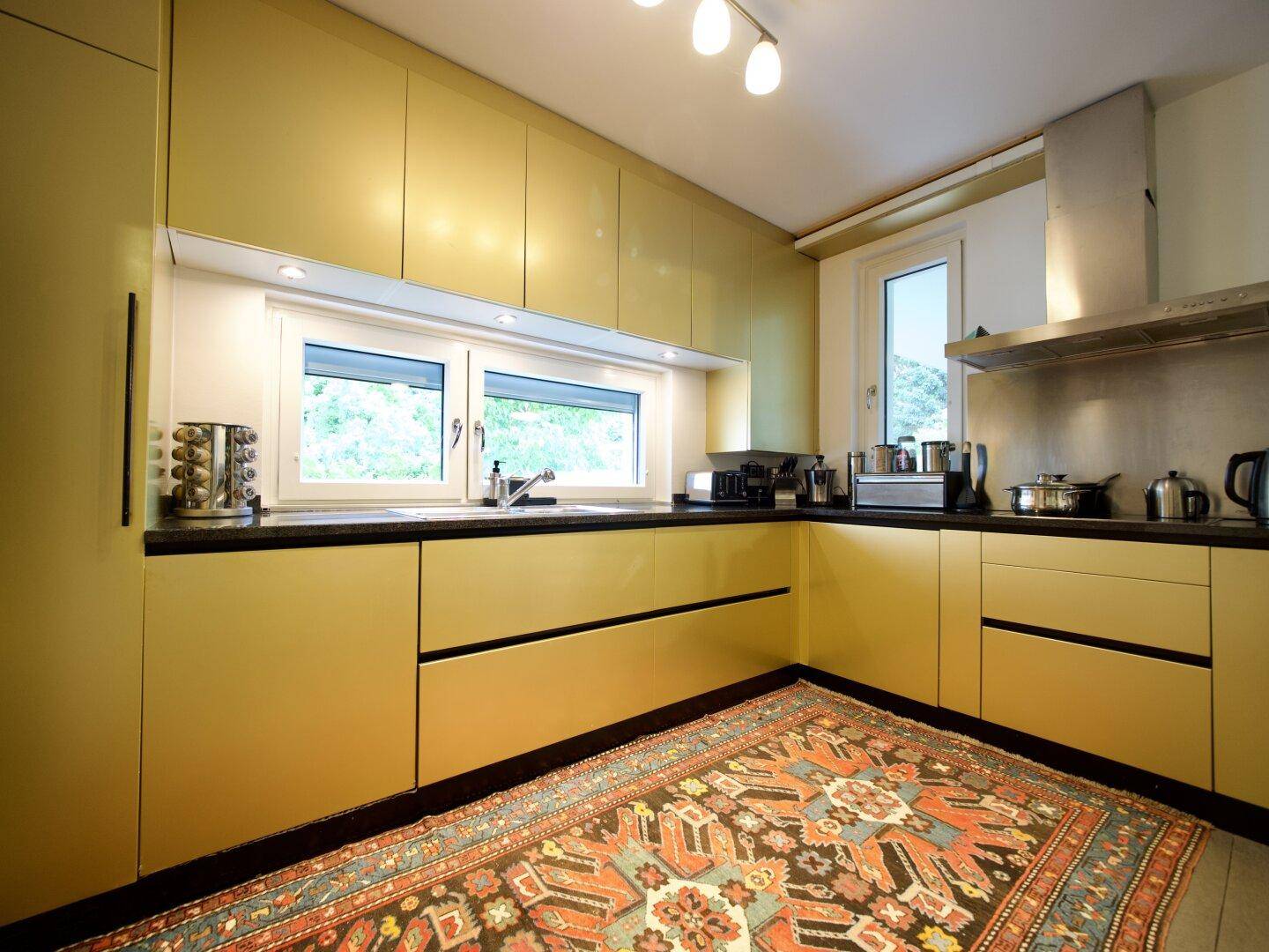 Moderne Küche mit glänzenden gelben Schränken, dunklen Arbeitsplatten und Fensterblick.