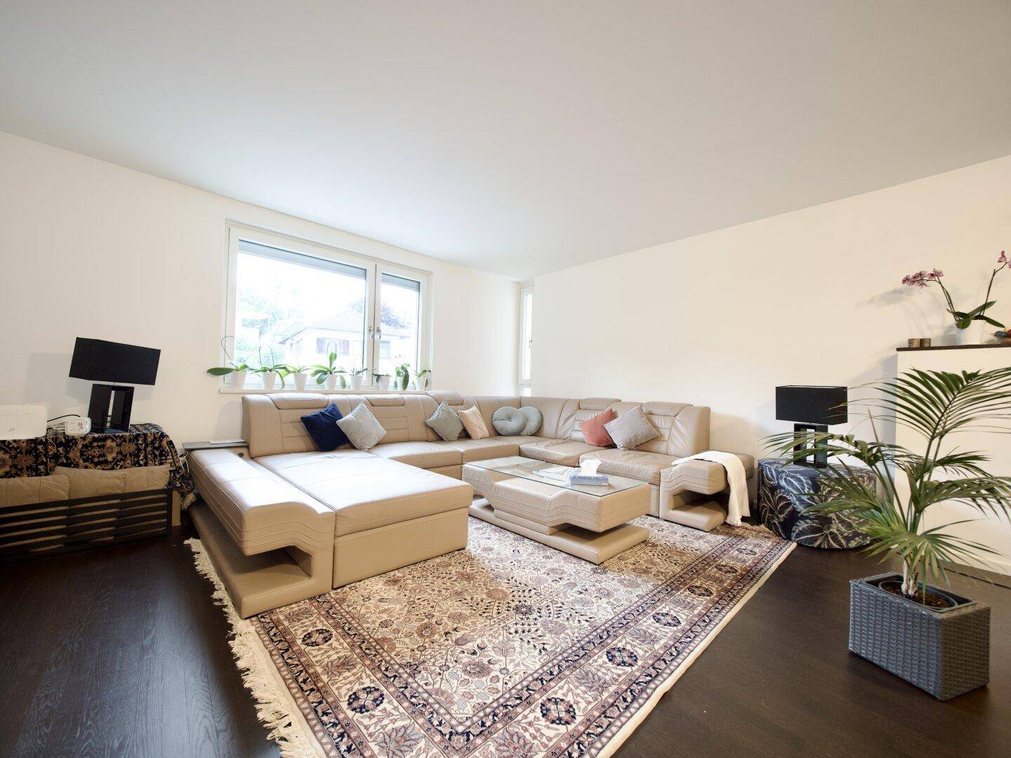 Heller Wohnbereich mit beigem Ecksofa, Couchtisch und einem Fenster mit Pflanzen.