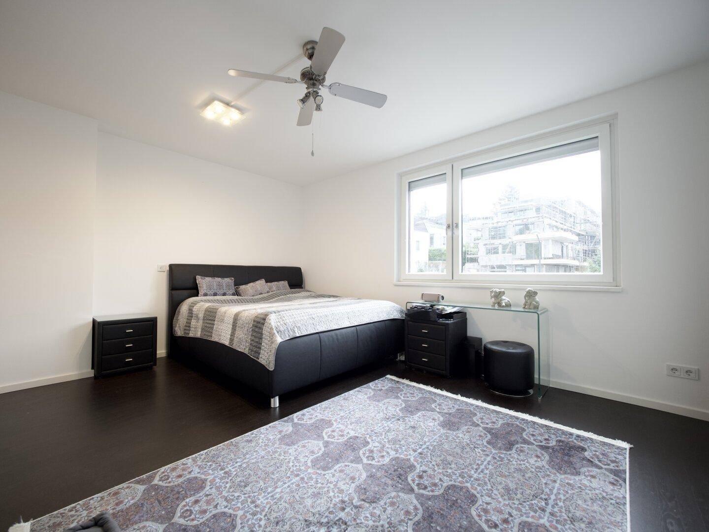 Helles Schlafzimmer mit großem Fenster, dunklem Bett und Deckenventilator.