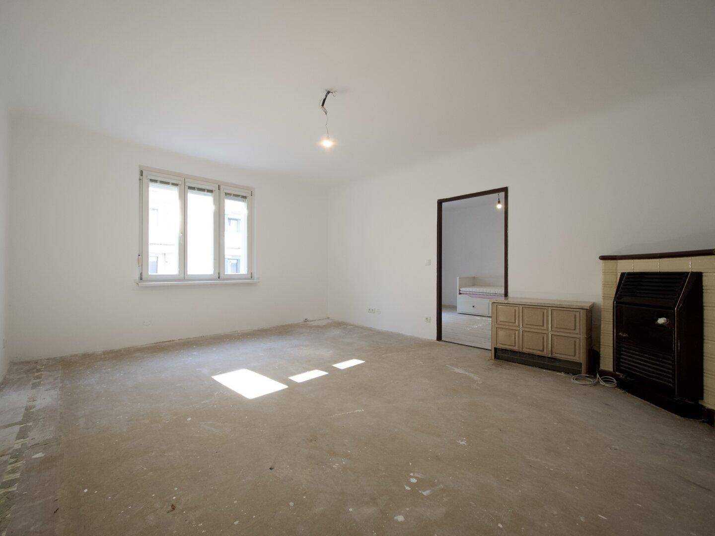Heller Raum mit Betonboden, Fenster und einem traditionellen Eckkamin.