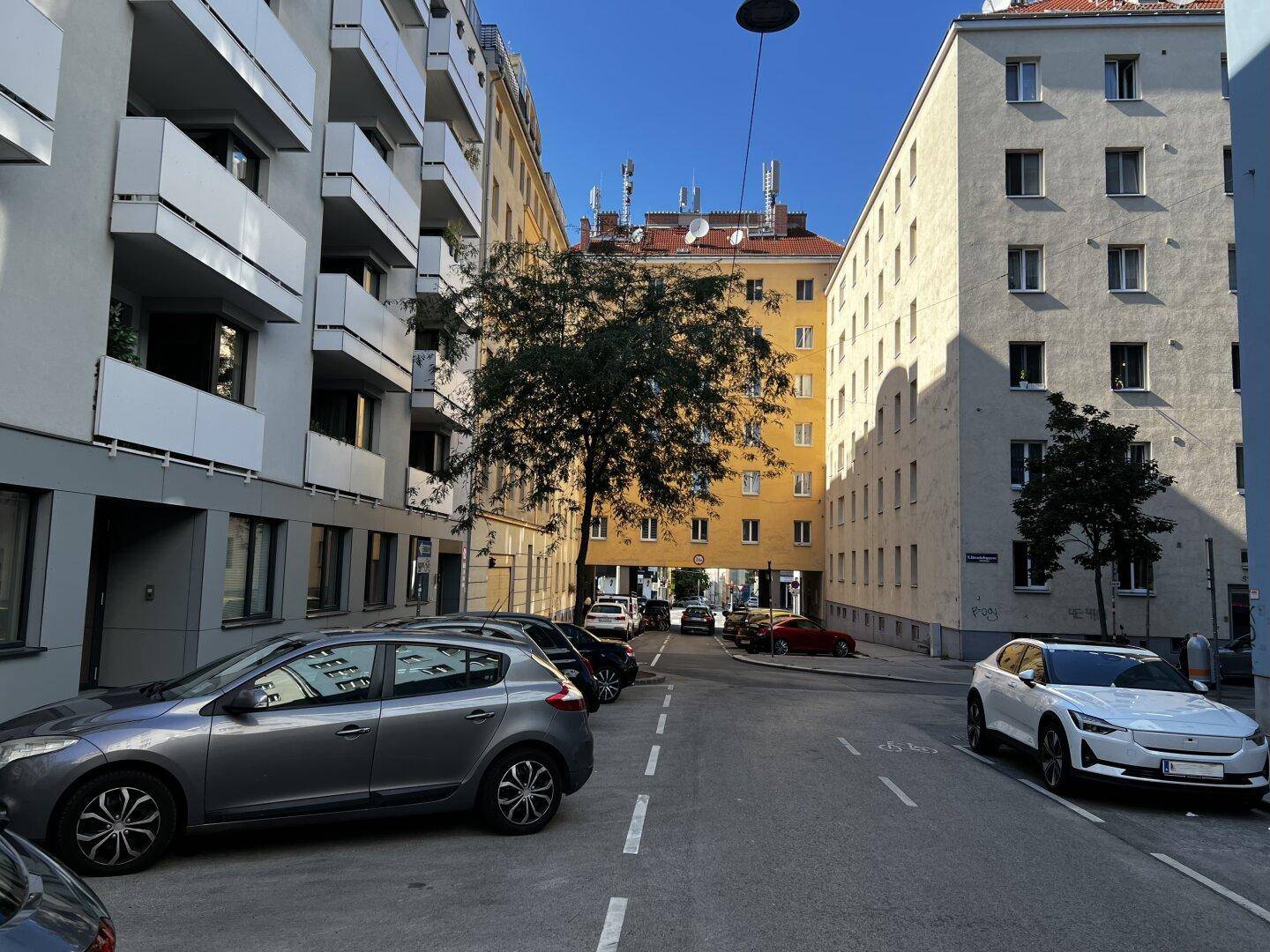 Städtische Straße mit Wohngebäuden, Bäumen und geparkten Fahrzeugen an einem sonnigen Tag.
