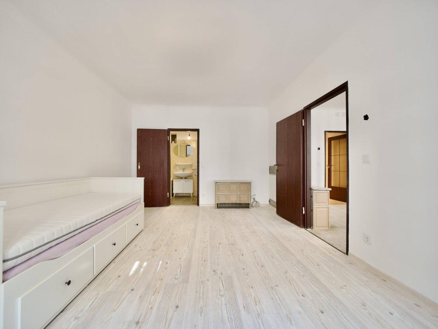 Heller Raum mit Holzboden und zwei Türen, eine davon führt zu einem Badezimmer.