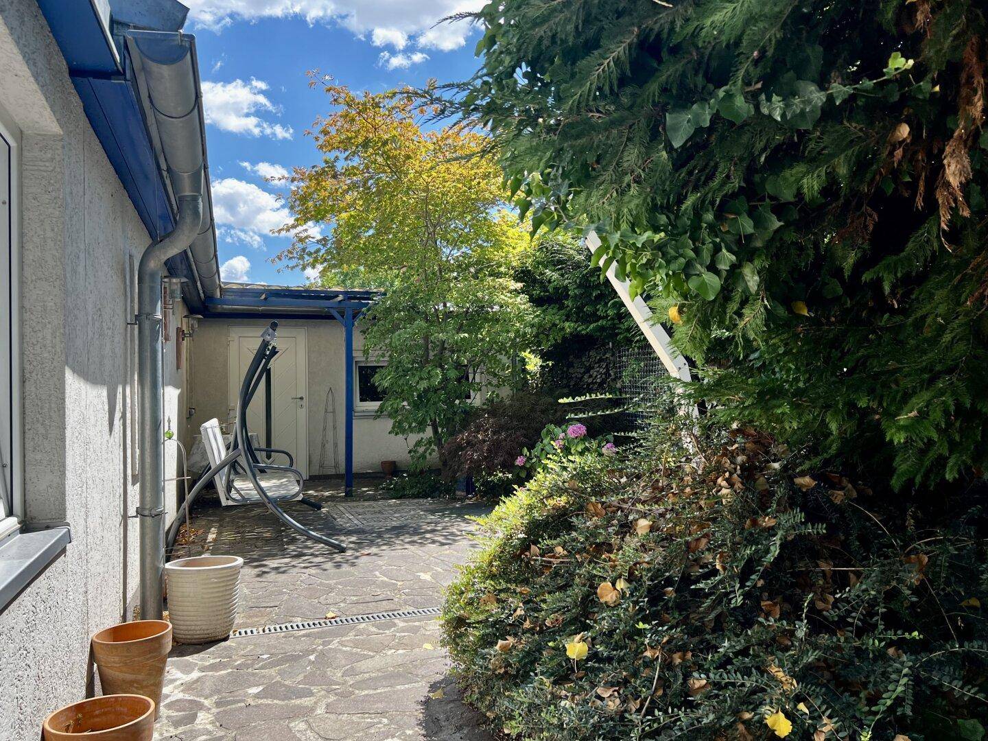 Eine sonnige Terrasse mit Schaukelbank und Topfpflanzen, umgeben von üppigem Grün.