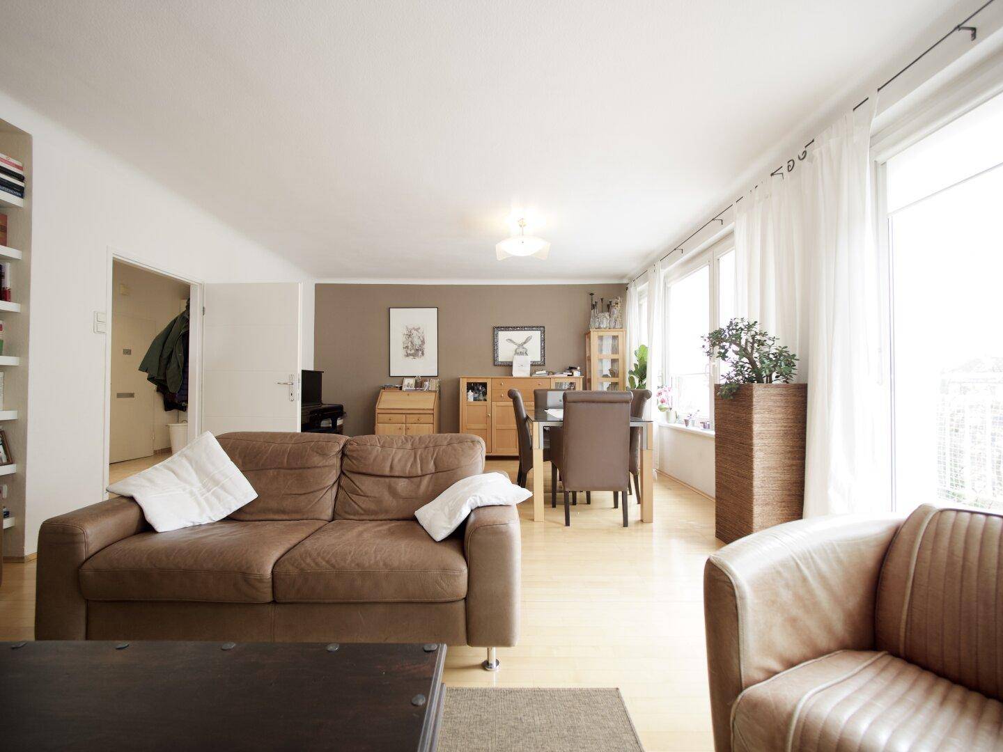 Gemütlicher Wohn-Essbereich mit braunem Sofa, Esstisch und viel Tageslicht durch große Fenster.