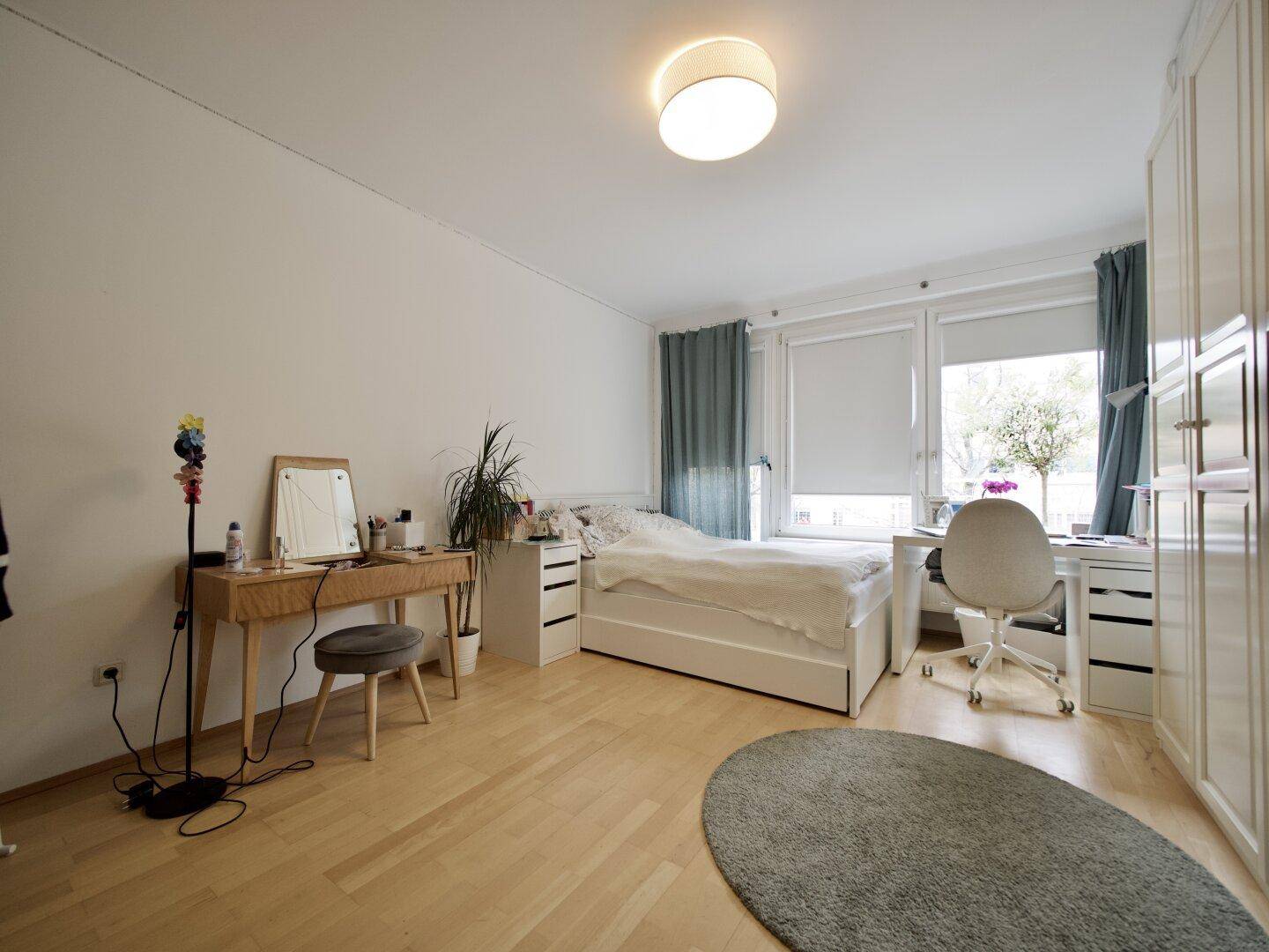 Helles Schlafzimmer mit Bett, Schreibtisch, Schminktisch und großen Fenstern.
