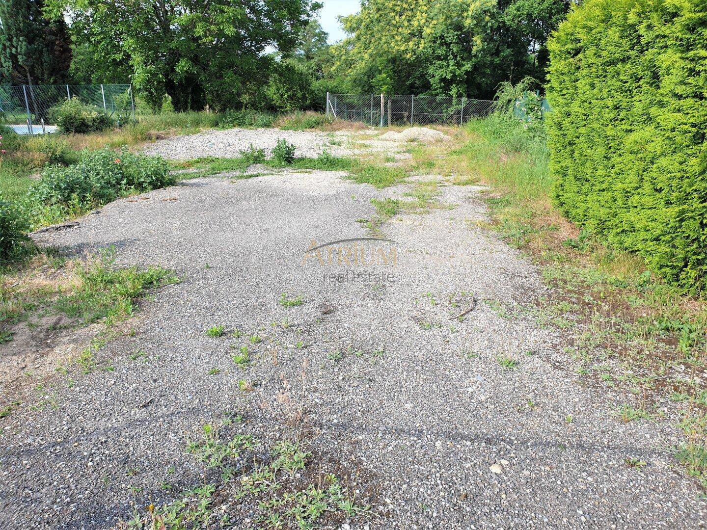 Schotterfläche auf einem Grundstück, umgeben von üppigem Grün und einer dichten Hecke.