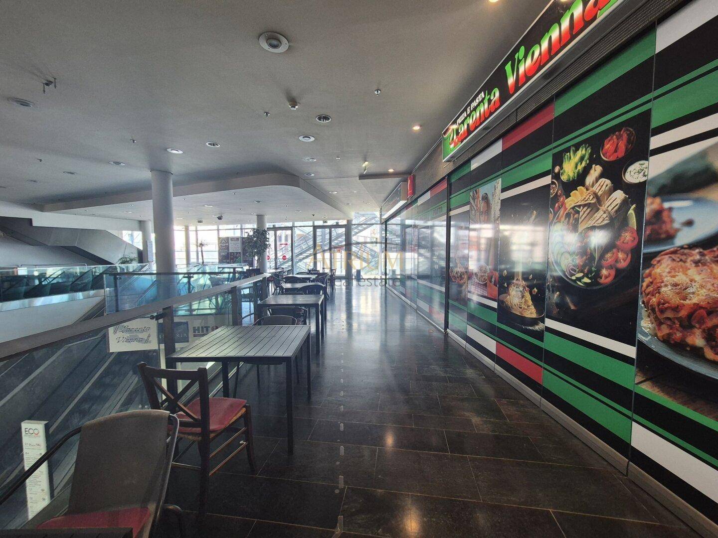 Langer Flur in einem Einkaufszentrum mit Restaurantfassaden und Sitzgelegenheiten entlang der Glasbrüstung.