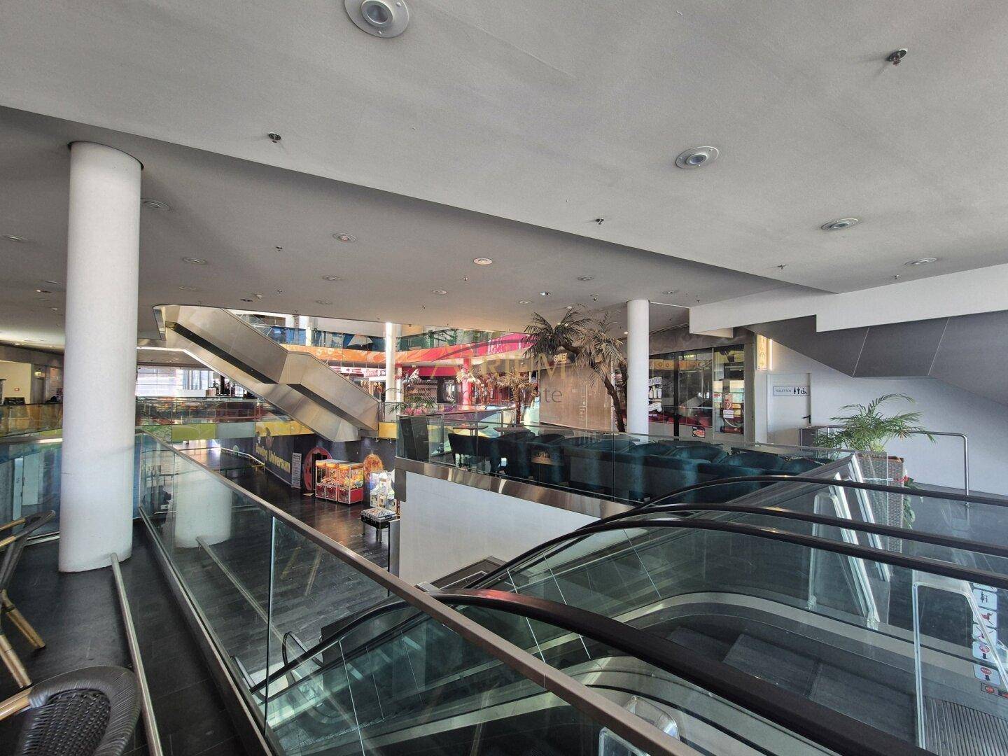Blick auf Rolltreppen und mehrere Ebenen in einem modernen Einkaufszentrum mit Sitzbereichen und Pflanzen.