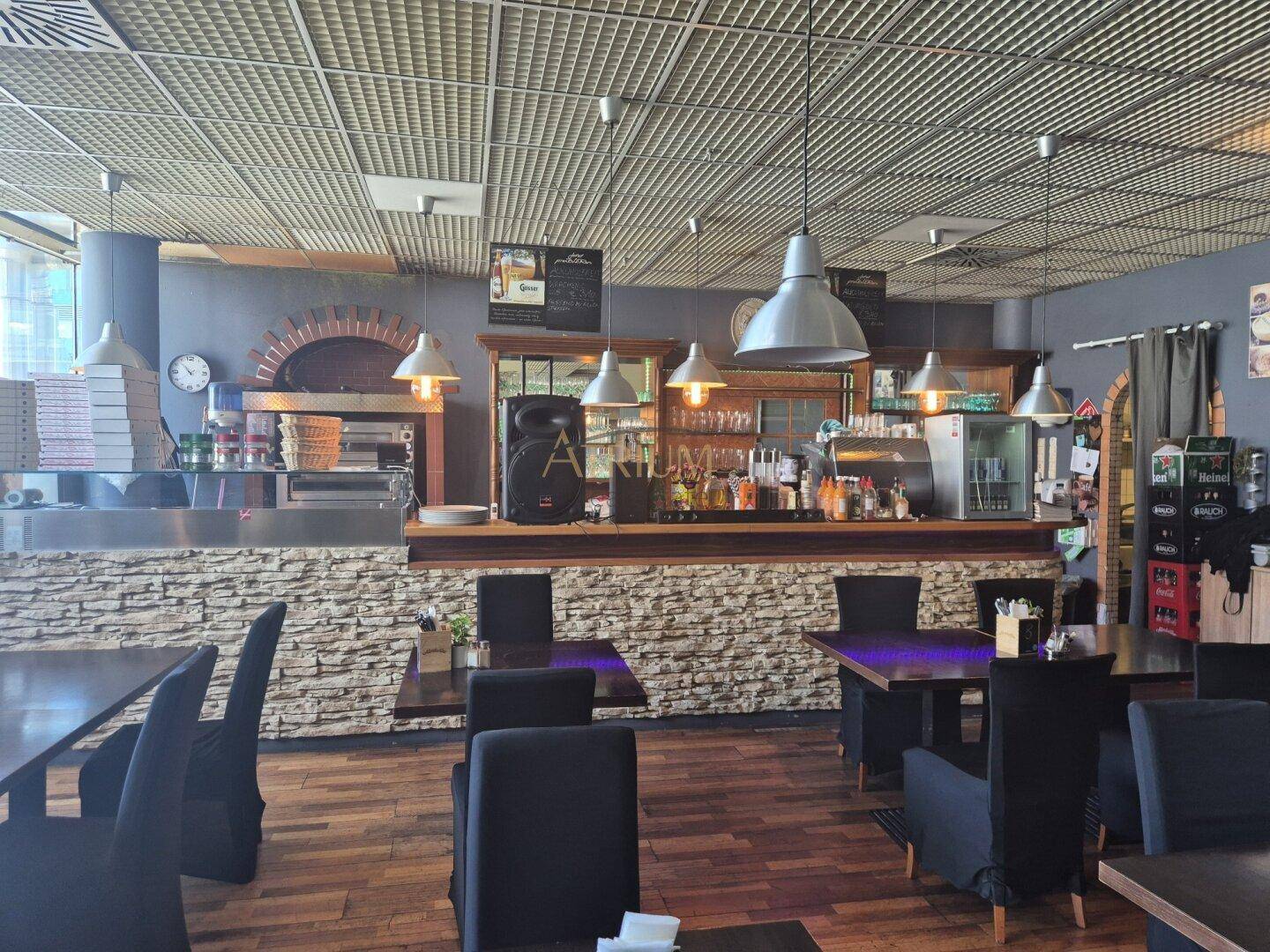 Innenansicht eines Restaurants mit Theke, Backofen und dunklen Tischen mit schwarzen Stühlen.