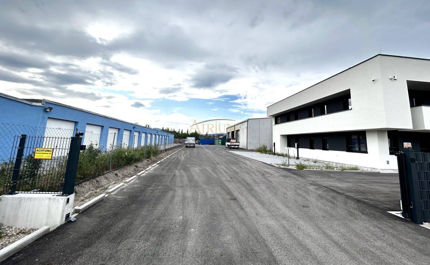 Außenansicht eines modernen Bürogebäudes neben einer Reihe blauer Lagerhallen unter bewölktem Himmel.