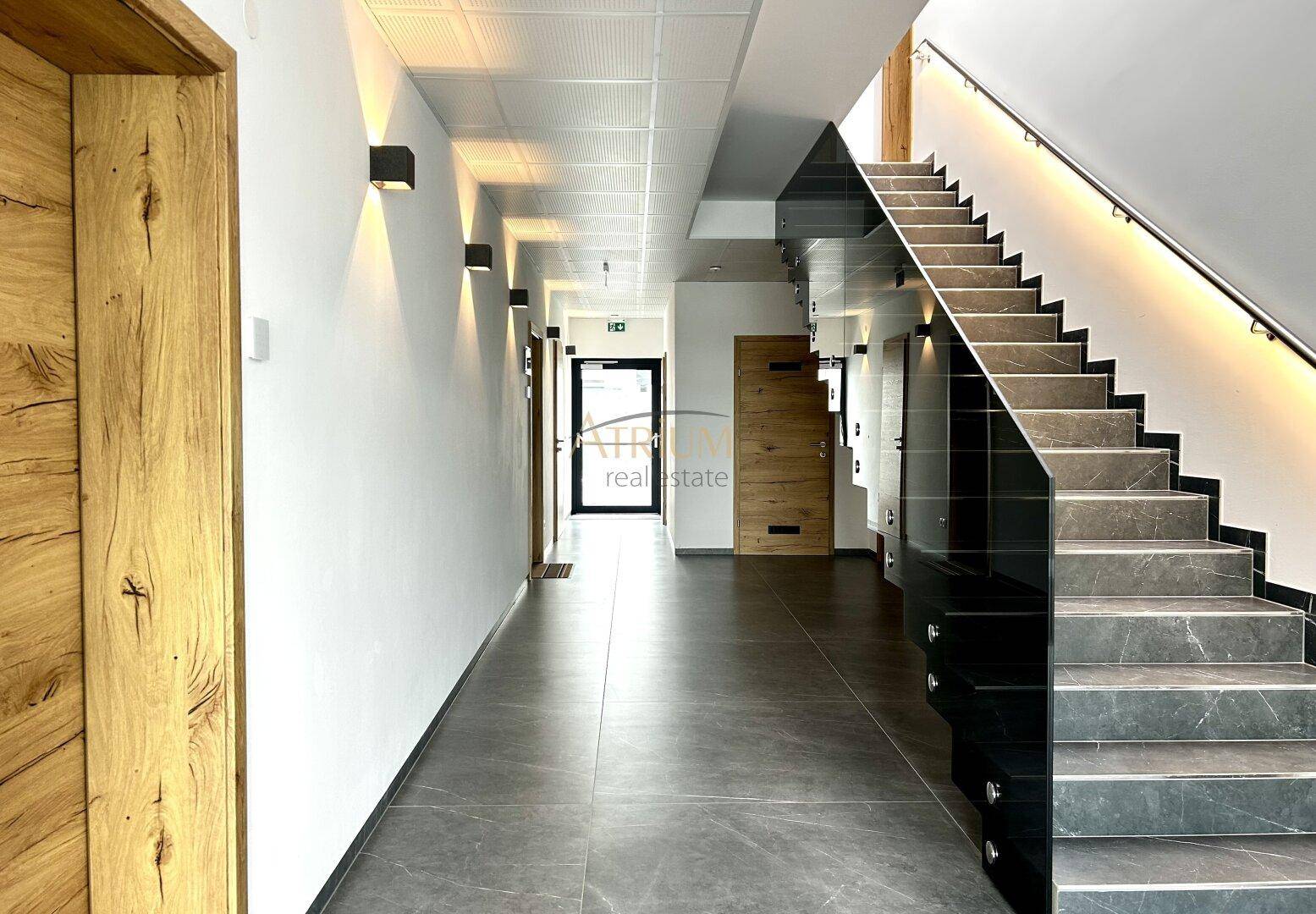 Heller Flur mit modernen Wandleuchten, Holzdetails und einer eleganten Treppe mit Glasgeländer.