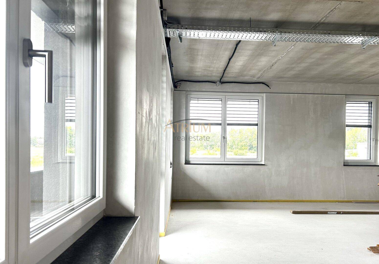 Unfertiger Büroraum mit großen Fenstern, Betonwänden und sichtbaren Deckeninstallationen.