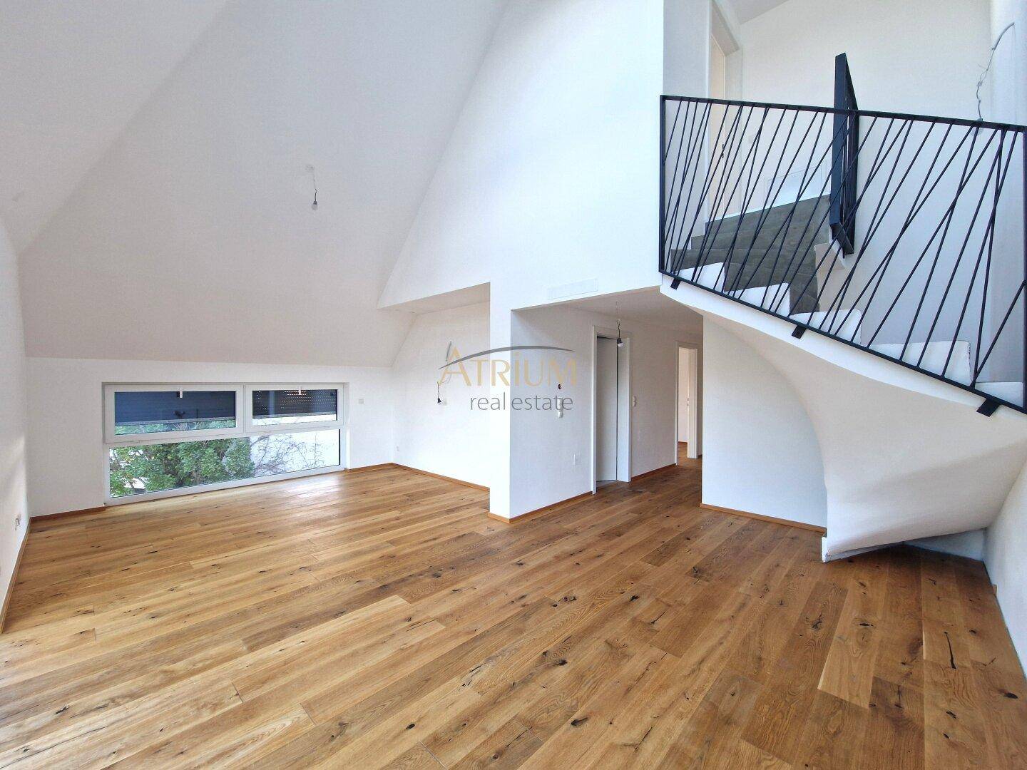 Heller Raum mit Holzboden, hohen Decken und einer modernen Treppe mit schwarzem Geländer.