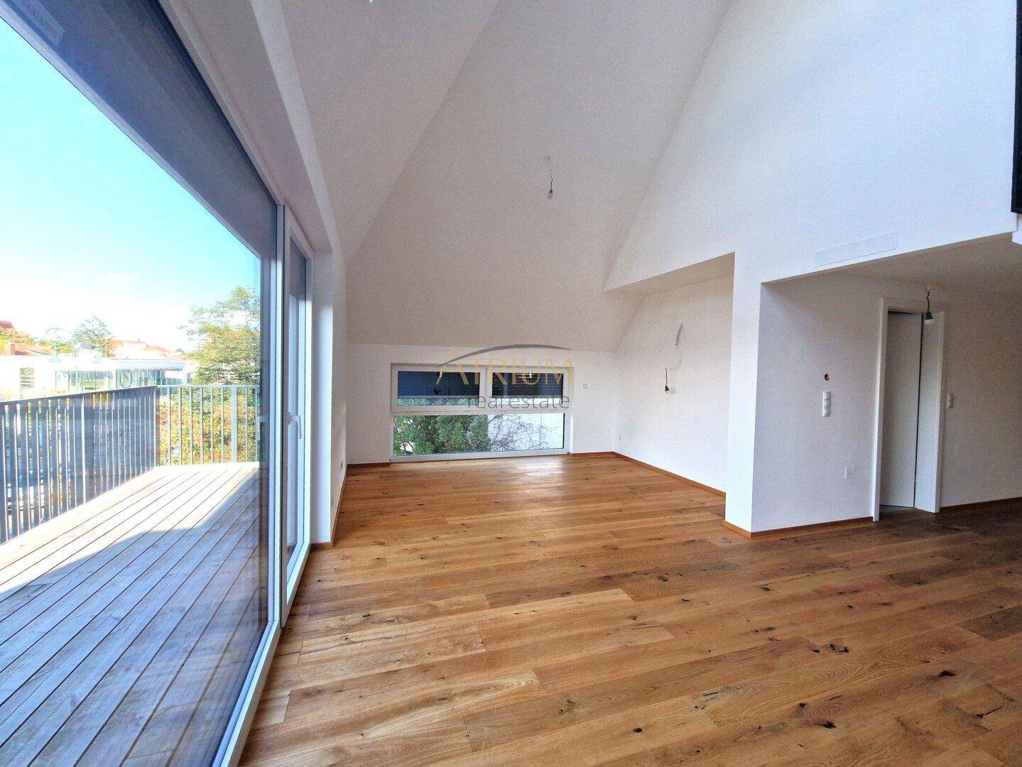 Heller, leerer Wohnbereich mit Holzboden, hohen Decken und großem Fenster mit Balkonzugang.
