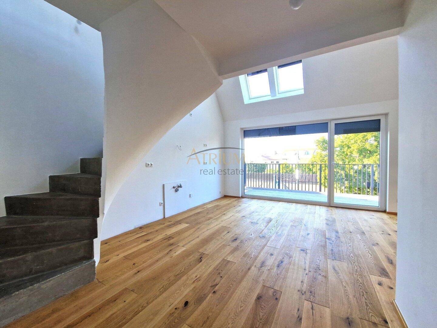 Geräumiger Raum mit Holzboden, Treppe, Balkonzugang und Dachfenstern, ideal für einen Wohnbereich.