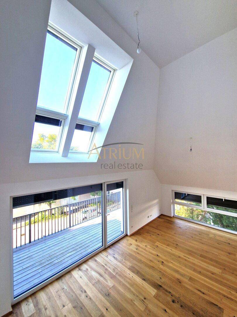 Lichtdurchfluteter Raum mit Holzboden, hohen Decken, großem Balkonfenster und mehreren Dachfenstern.