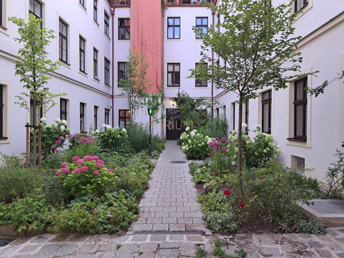Idyllischer Innenhofgarten mit gepflastertem Weg, blühenden Pflanzen und jungen Bäumen.