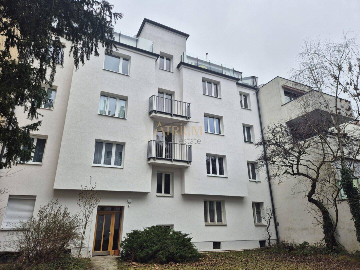 Mehrfamilienhaus mit weißer Fassade, Balkonen und großen Fenstern in grüner Umgebung.