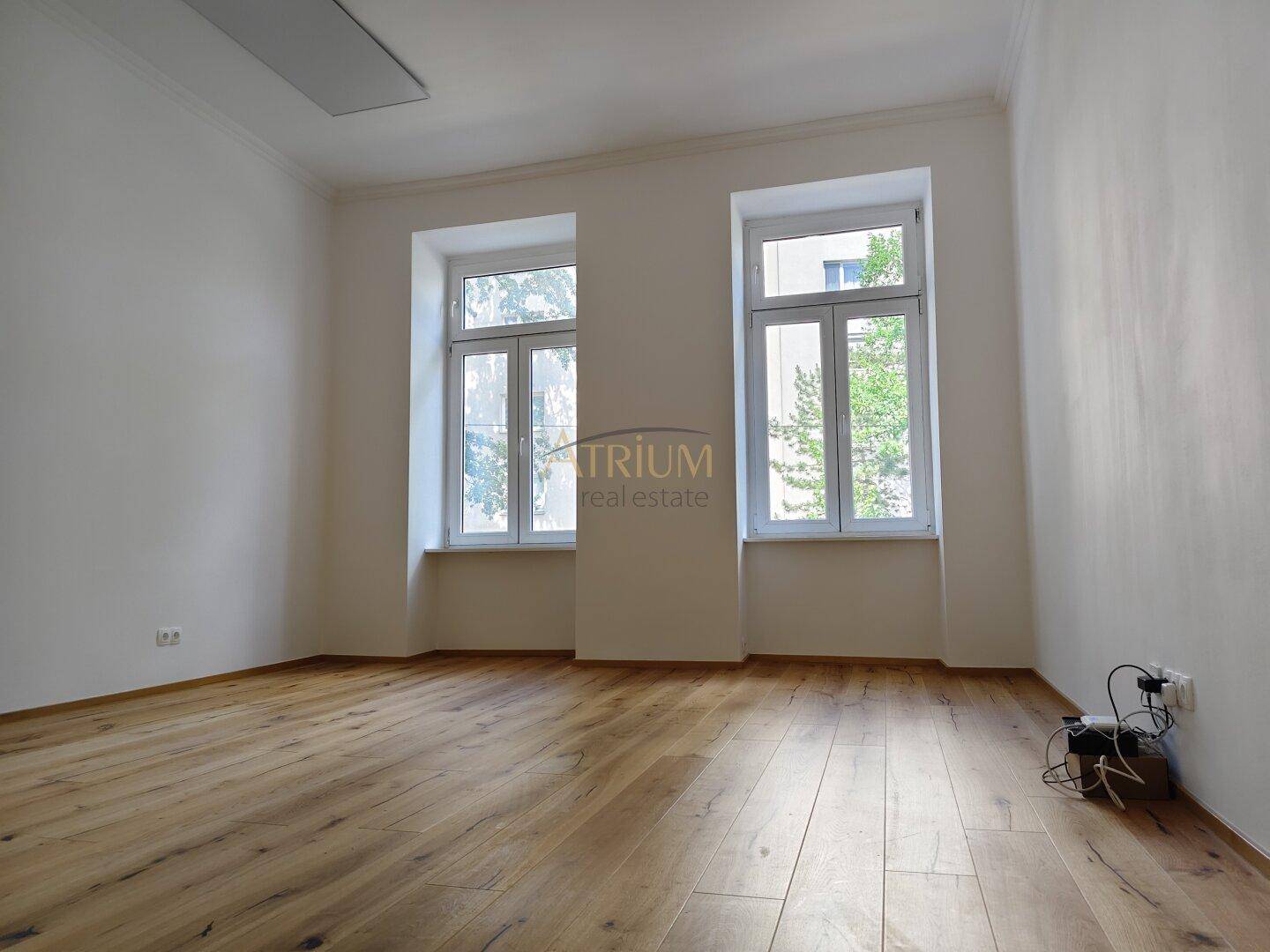Helles, leeres Zimmer mit Holzboden und zwei großen Fenstern, die viel Tageslicht hereinlassen.