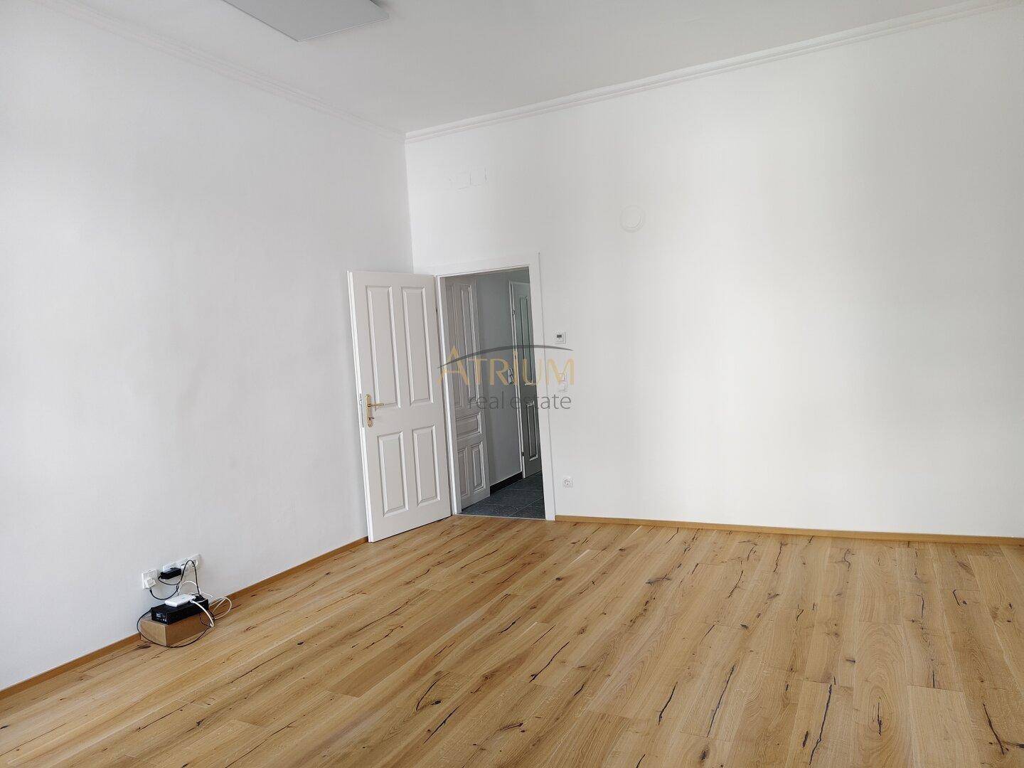Geräumiges, leeres Zimmer mit Holzboden und einer offenen Tür, die zu einem weiteren Raum führt.
