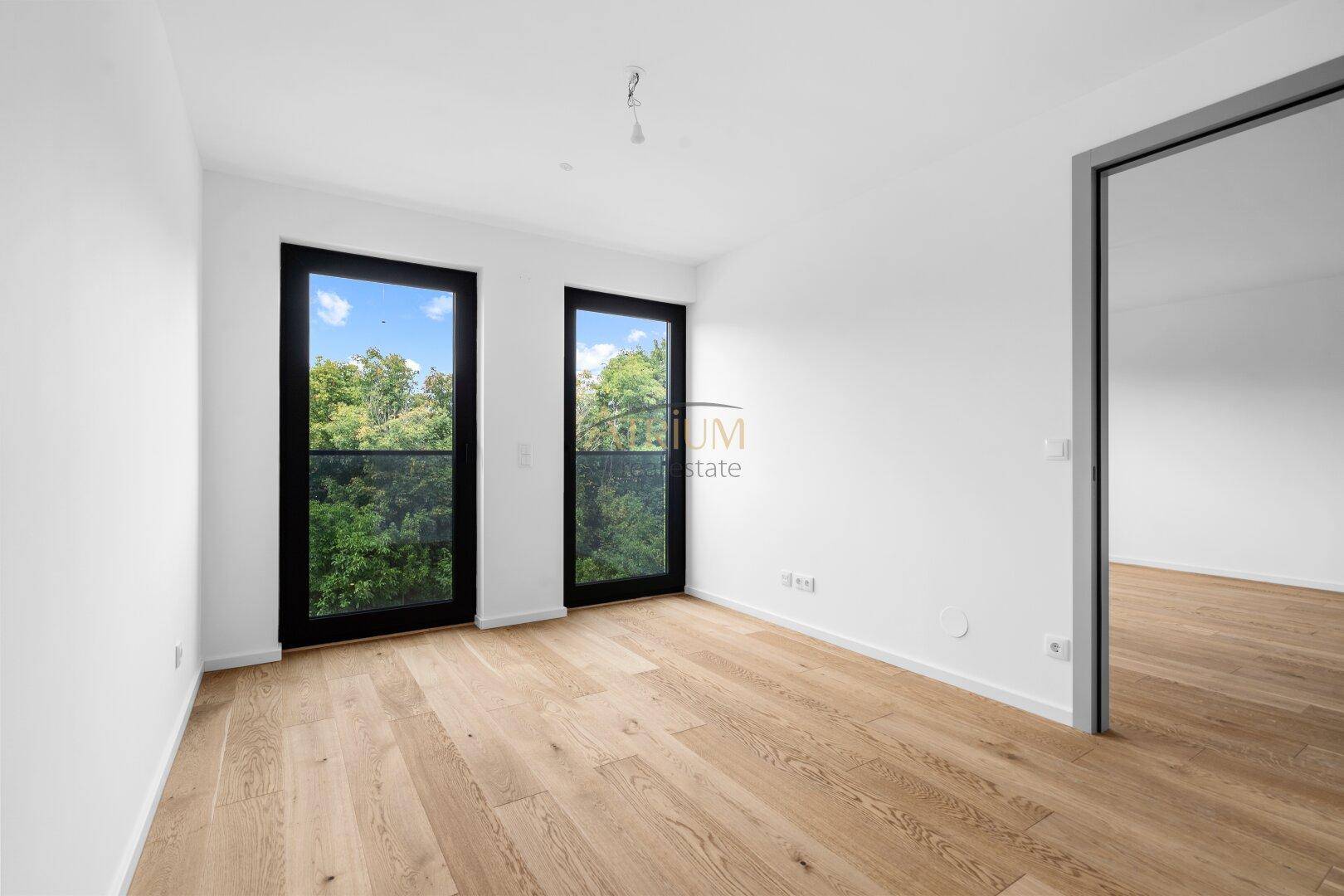 Geräumiger, heller Raum mit Holzboden und bodentiefen Fenstern, die viel Tageslicht hereinlassen.