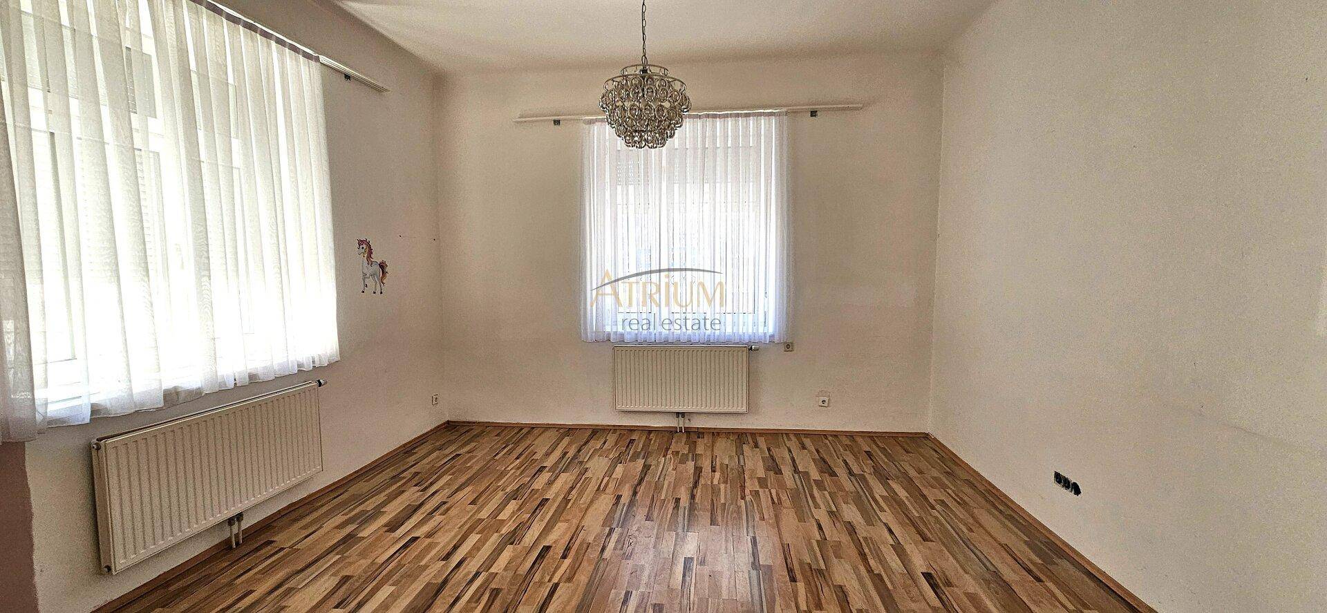 Heller Raum mit Holzboden, großem Fenster und einem Kronleuchter an der Decke.