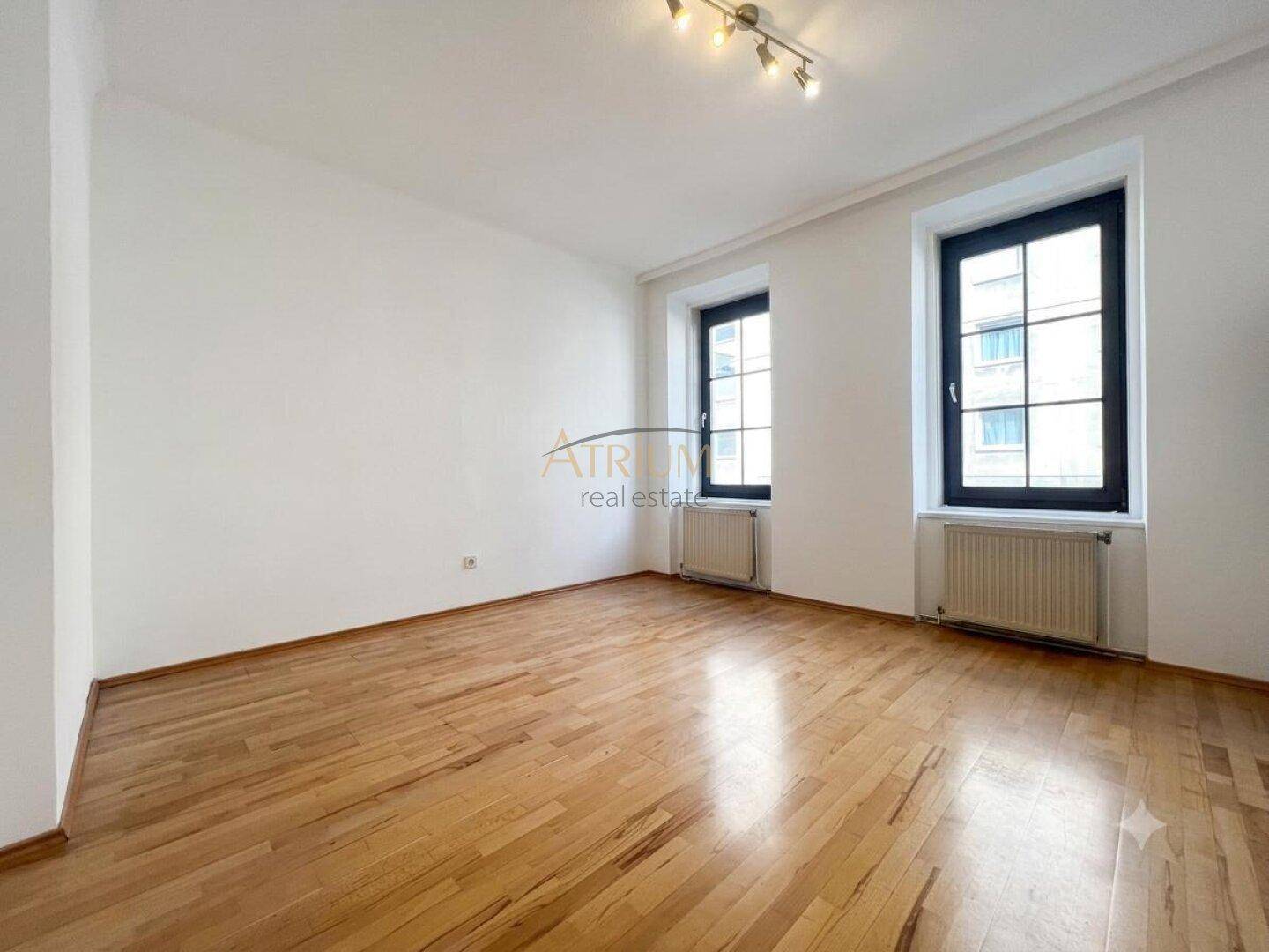 Heller Raum mit zwei Fenstern, Holzfußboden und weißen Wänden, bietet viel Tageslicht.