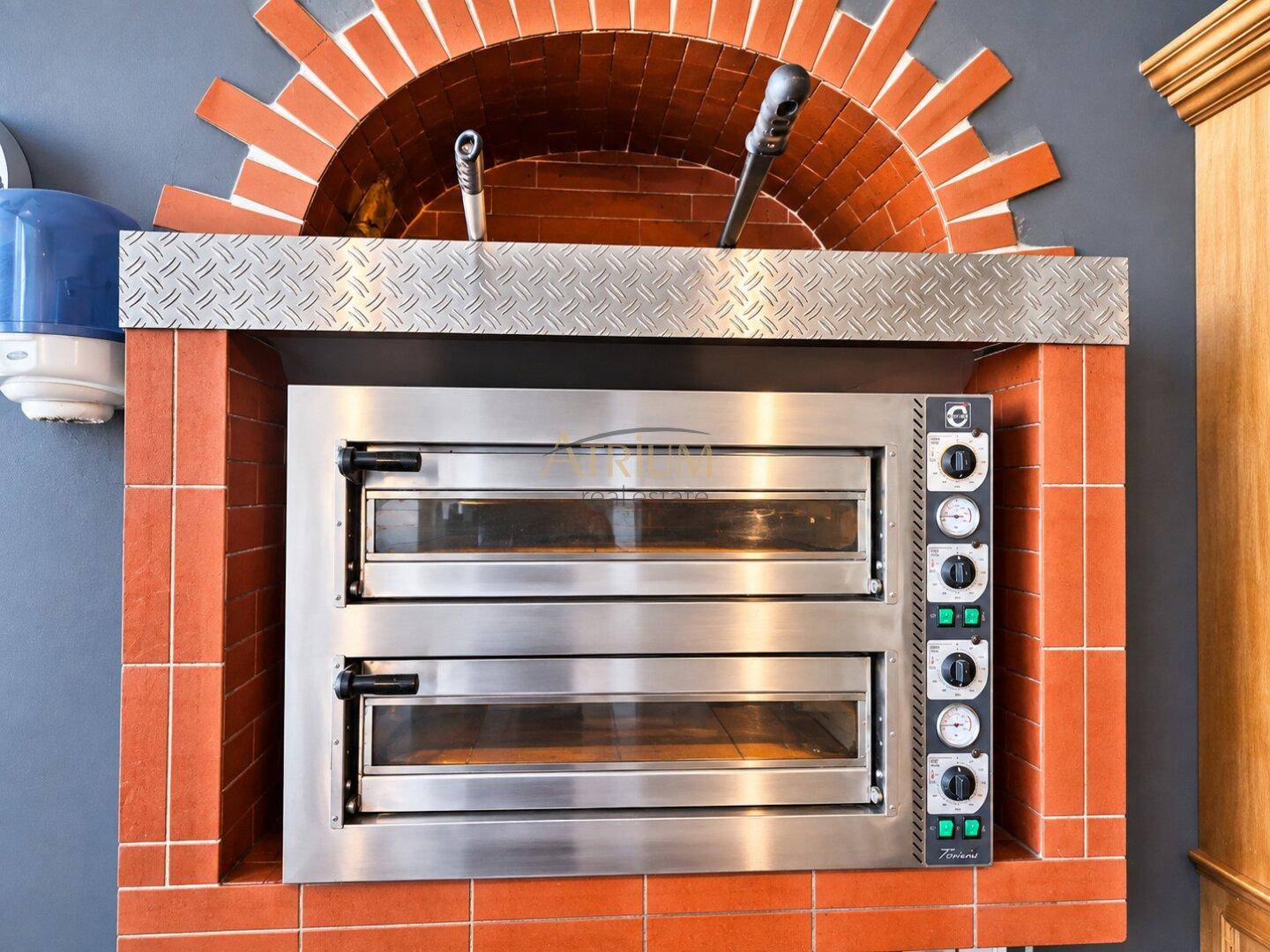 Professioneller Pizzaofen mit zwei Kammern, umgeben von roten Ziegeln und Edelstahlverkleidung in einer Restaurantküche.
