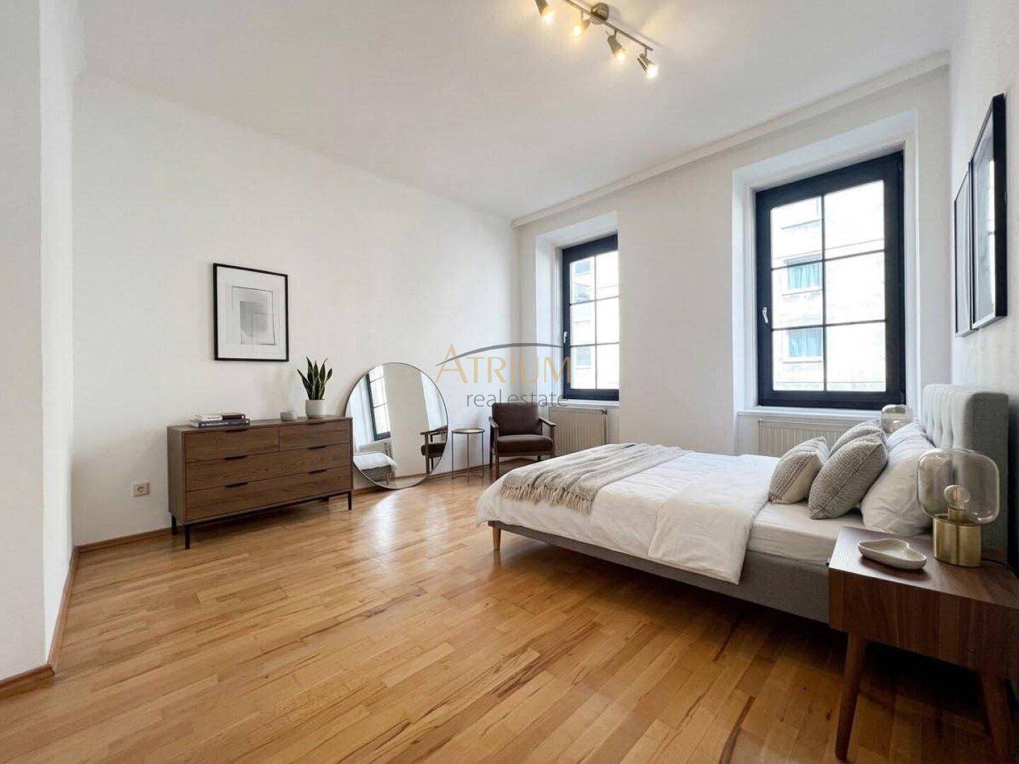 Geräumiges Schlafzimmer mit Holzboden, großem Bett, Kommode und natürlichem Licht durch die Fenster.