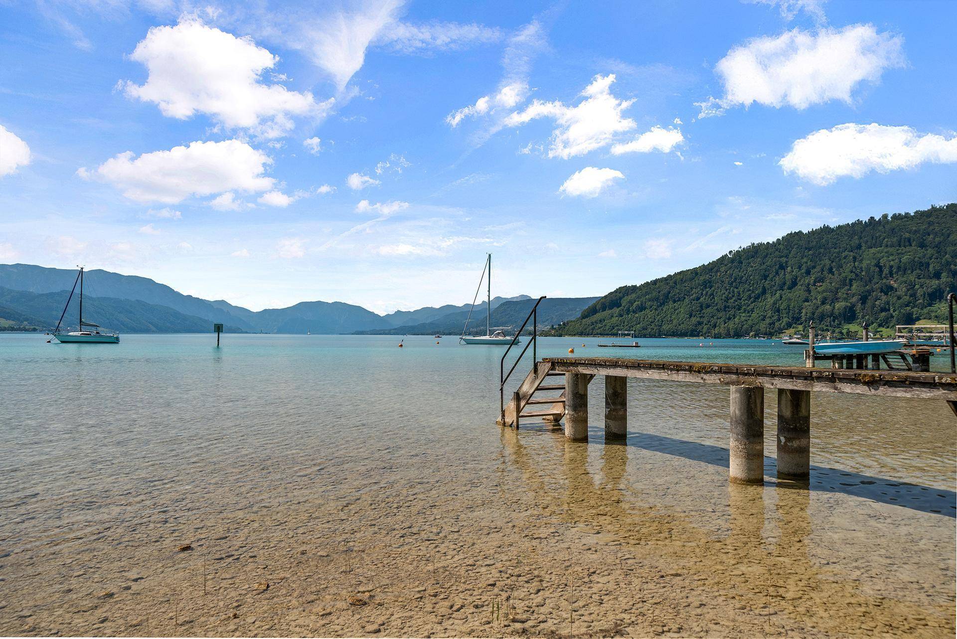 Betonsteg ragt ins klare Seewasser, umgeben von Segelbooten und grünen Berghängen.