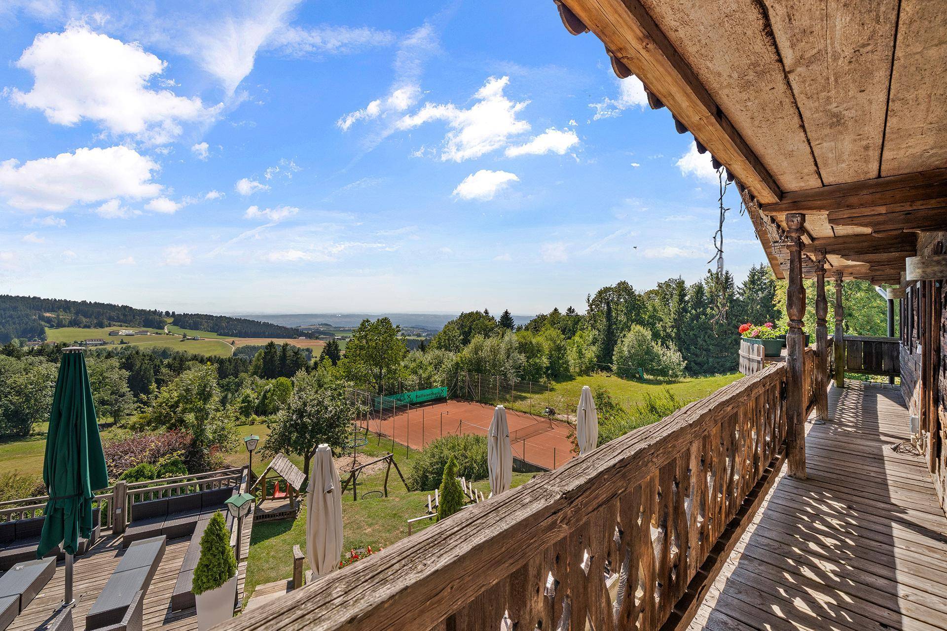 Langer Holzbalkon mit Blick auf die weite Landschaft, Tennisplatz und grüne Umgebung.
