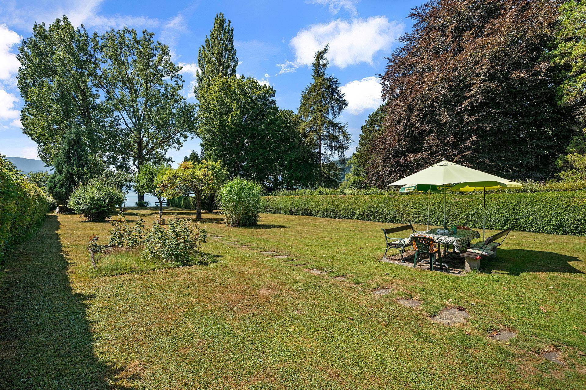 Idyllischer Gartenbereich mit Rasen, Blumenbeeten und einer einladenden Sitzgruppe unter einem Sonnenschirm.