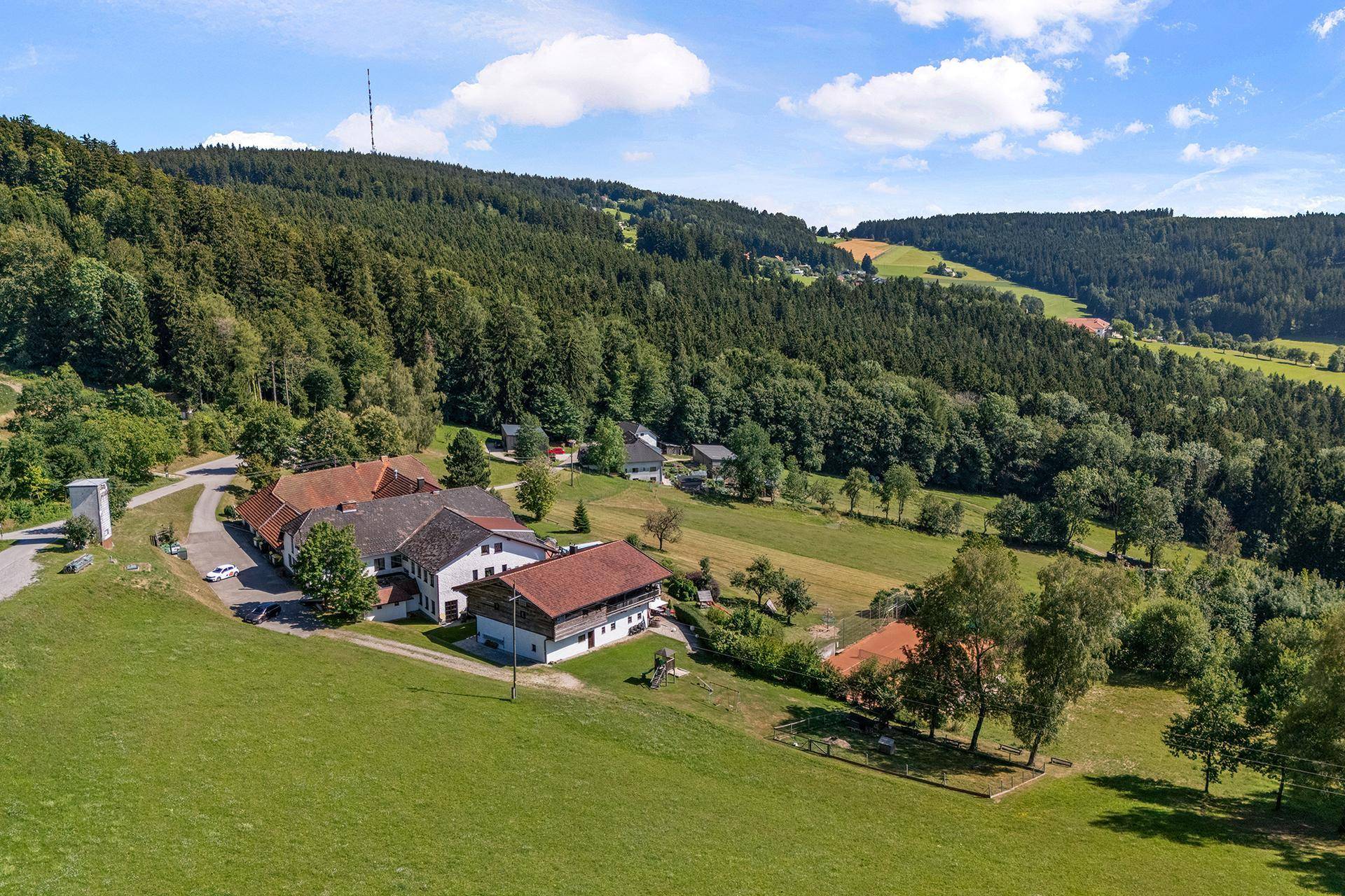 Luftaufnahme des Anwesens eingebettet in eine grüne, bewaldete Hügellandschaft unter blauem Himmel.