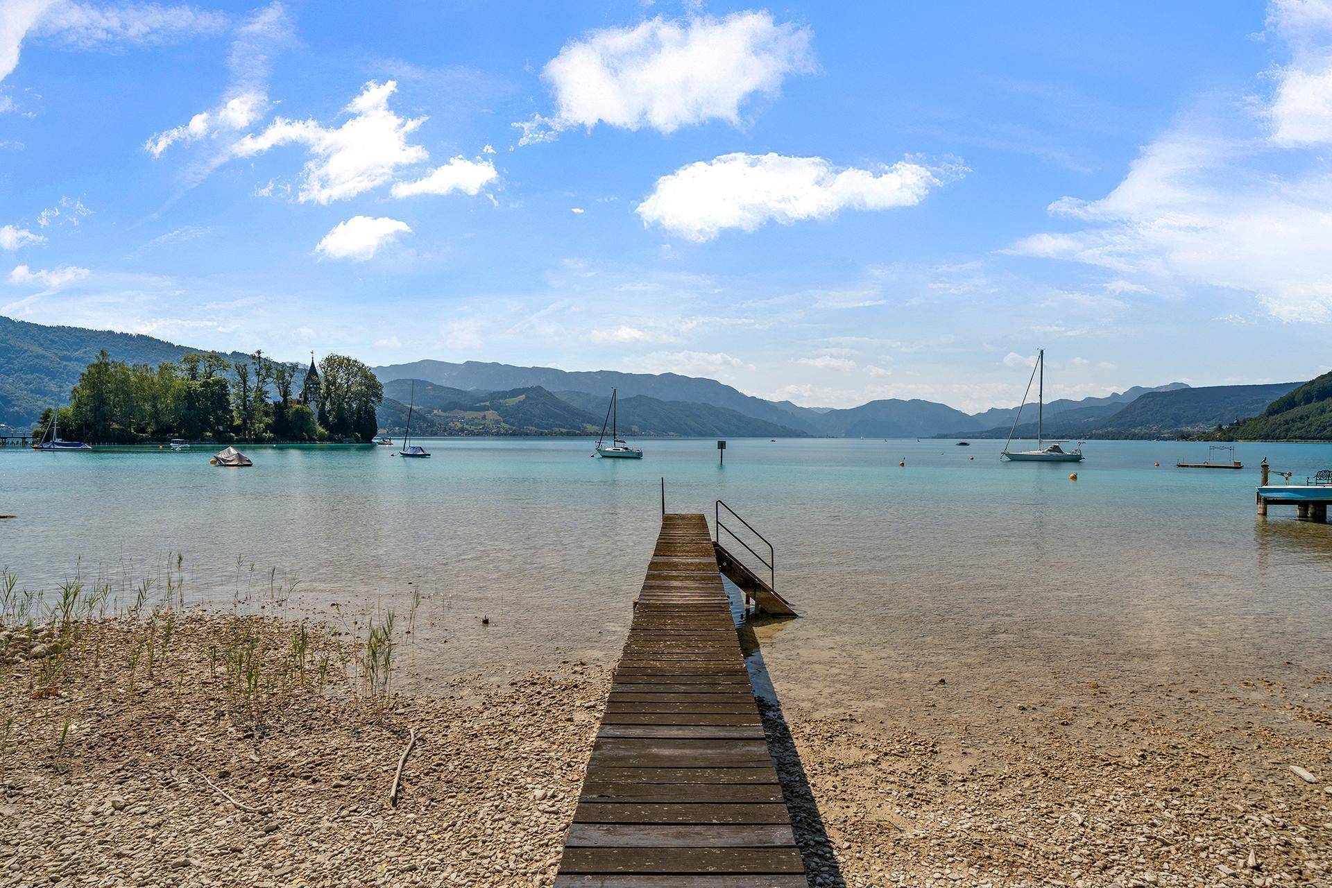 Langer Holzsteg führt ins klare Seewasser mit Segelbooten und Bergpanorama im Hintergrund.
