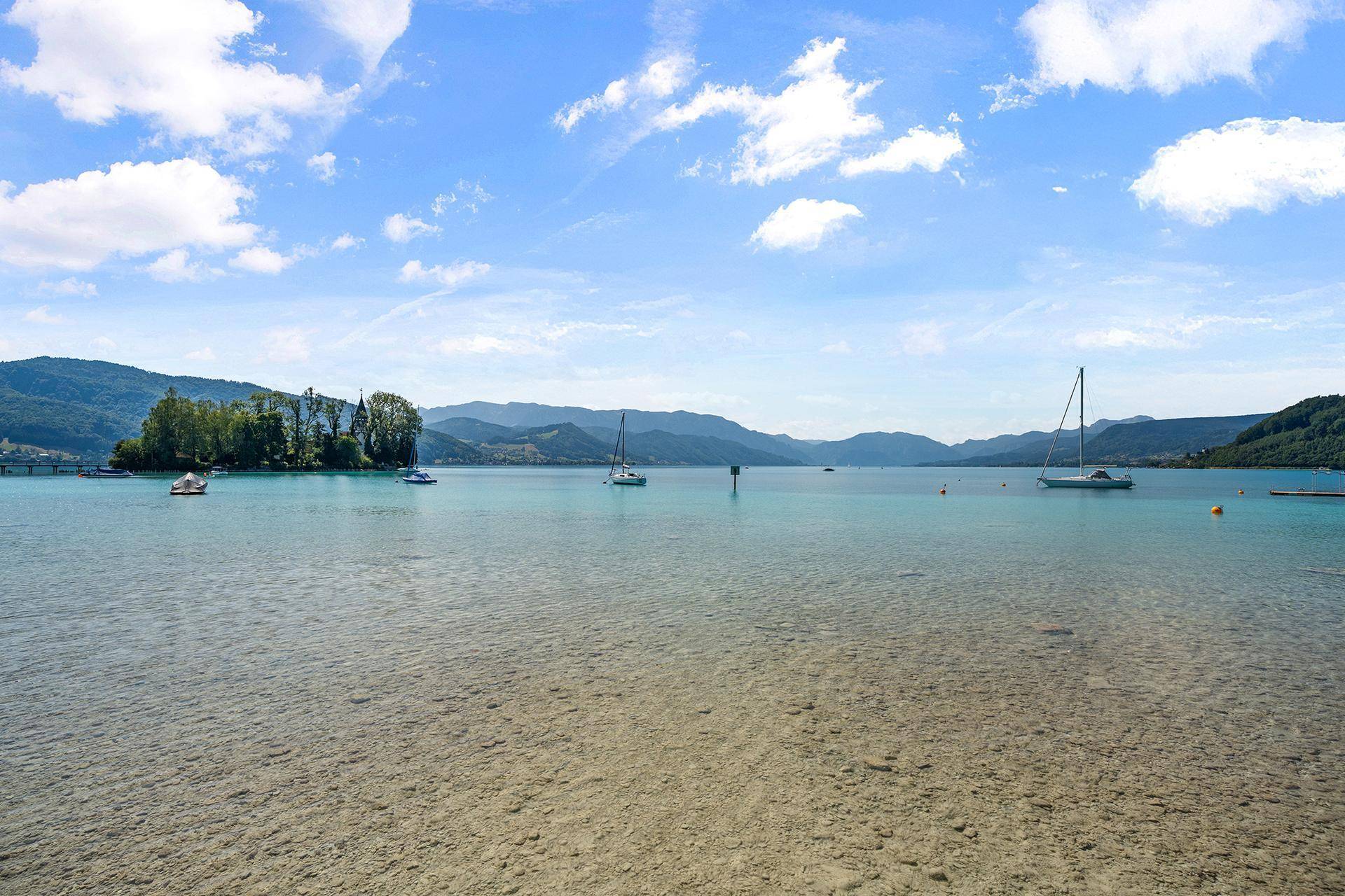 Weitläufiger Blick über den ruhigen See mit Segelbooten und bewaldeten Bergen am Horizont.