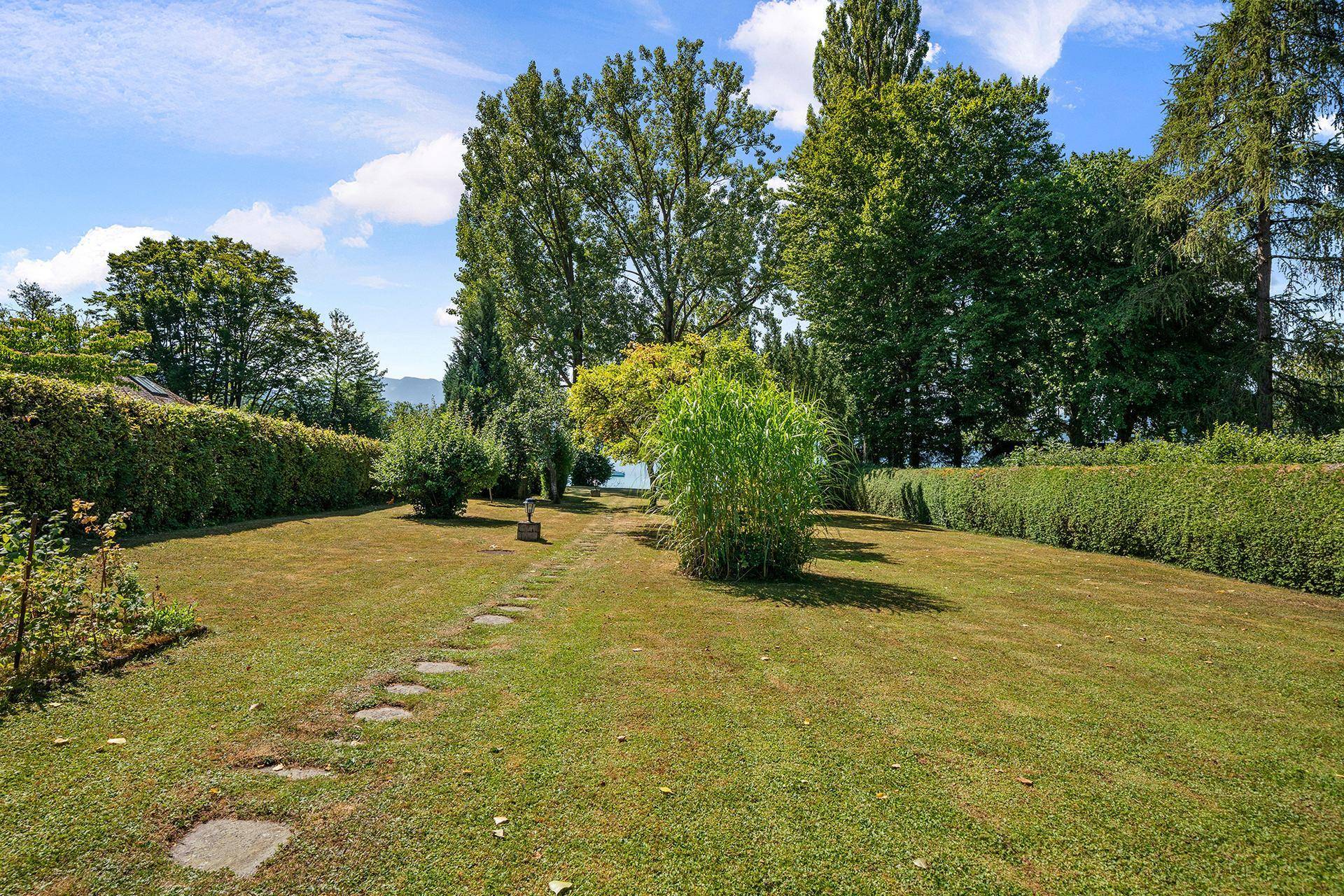 Weitläufiger Rasengarten mit Steinplattenweg, hohen Hecken und Blick auf den See im Hintergrund.