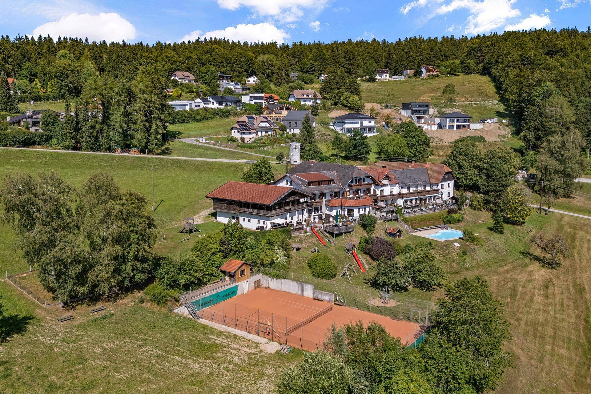 Übersichtliche Luftaufnahme des Anwesens mit Tennisplatz, Pool und Spielplatz in idyllischer Natur.