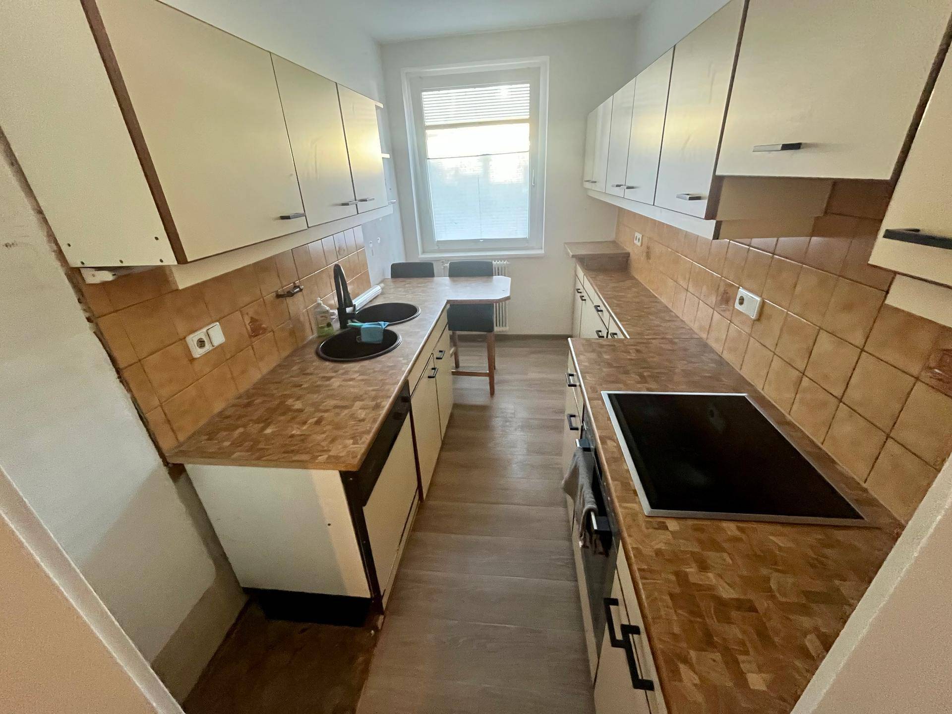 Funktionale Küche mit beigen Fliesen, Holzoptik-Arbeitsplatten und Fenster für Tageslicht.