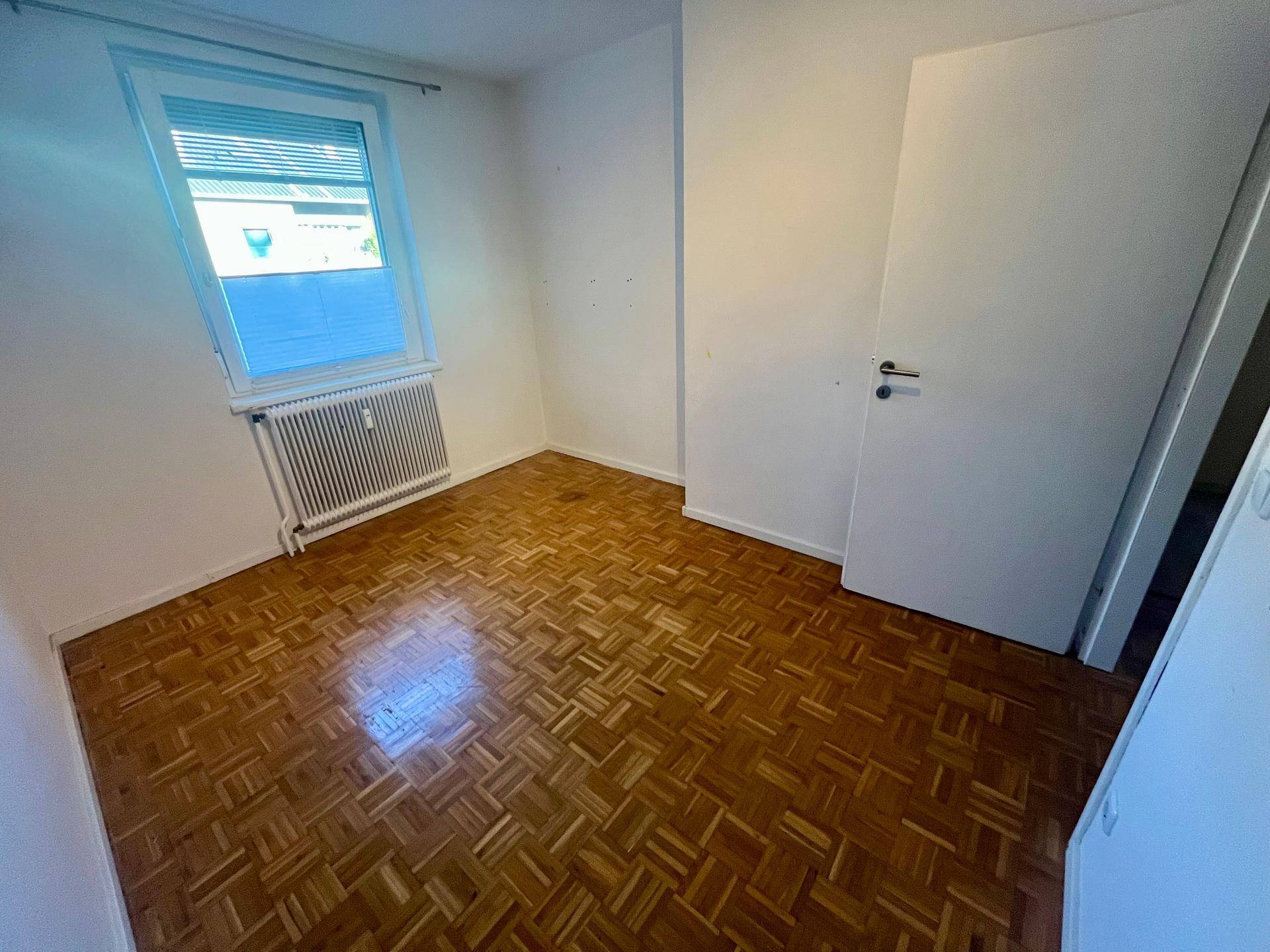 Schlafzimmer mit Parkettboden, Fenster und weißer Tür, bereit zur individuellen Gestaltung.