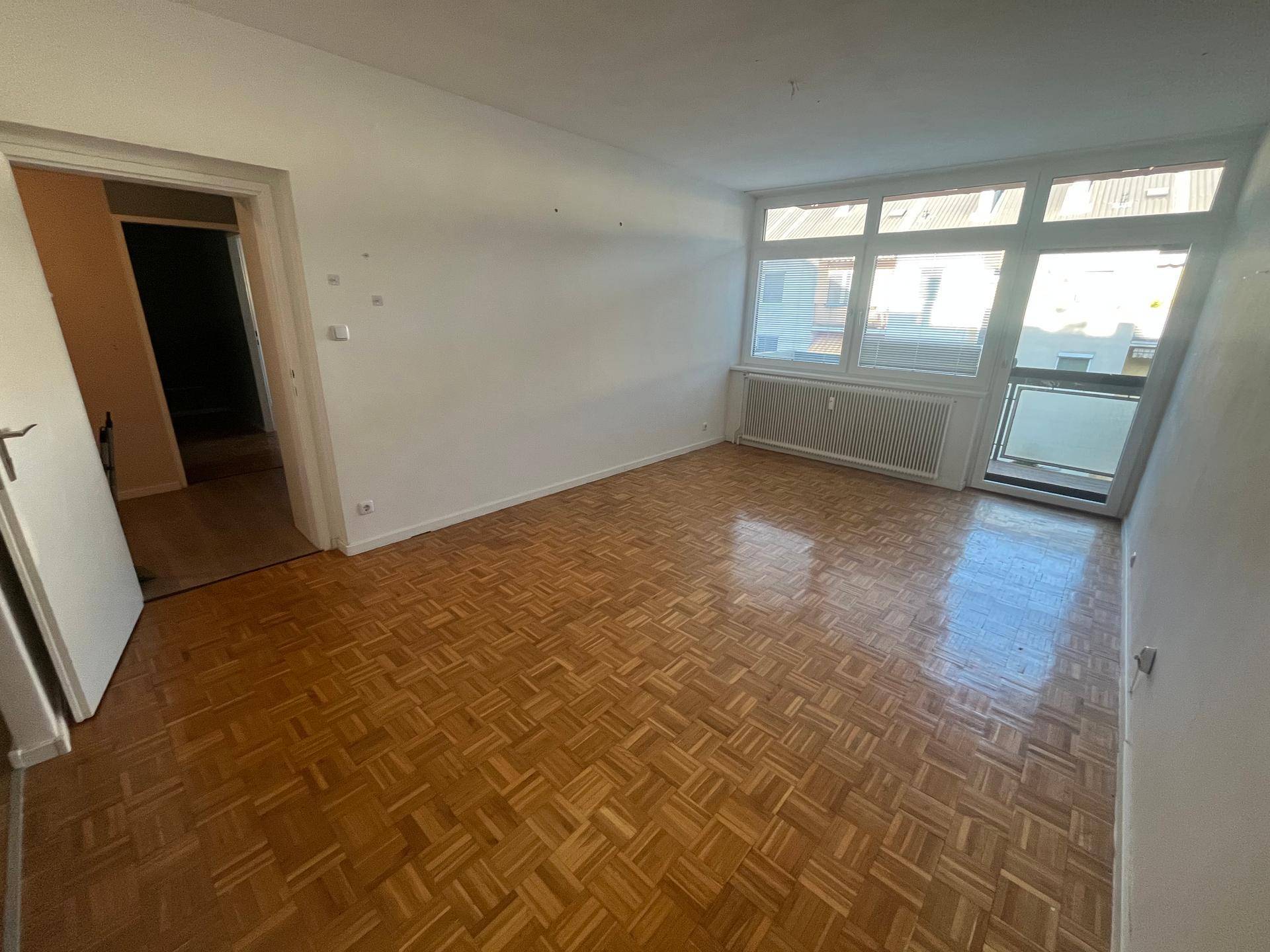 Geräumiges Wohnzimmer mit großen Fenstern, Parkettboden und Heizkörper unter dem Fenster.