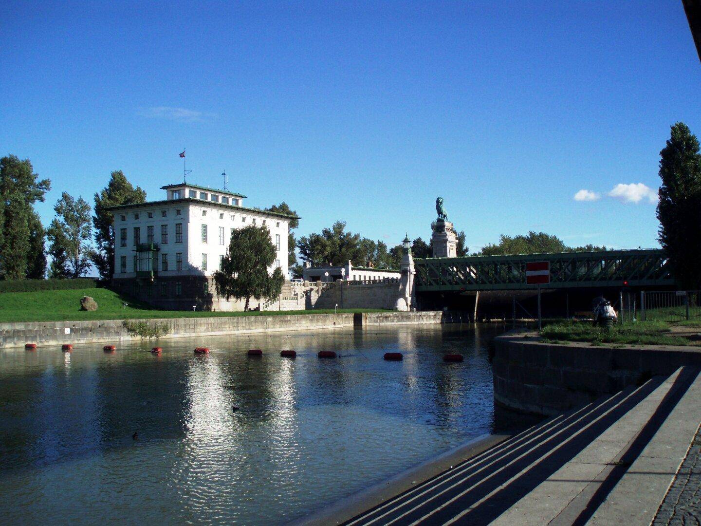 Malerischer Flusslauf mit einem historischen weißen Gebäude und einer Brücke unter klarem blauem Himmel.