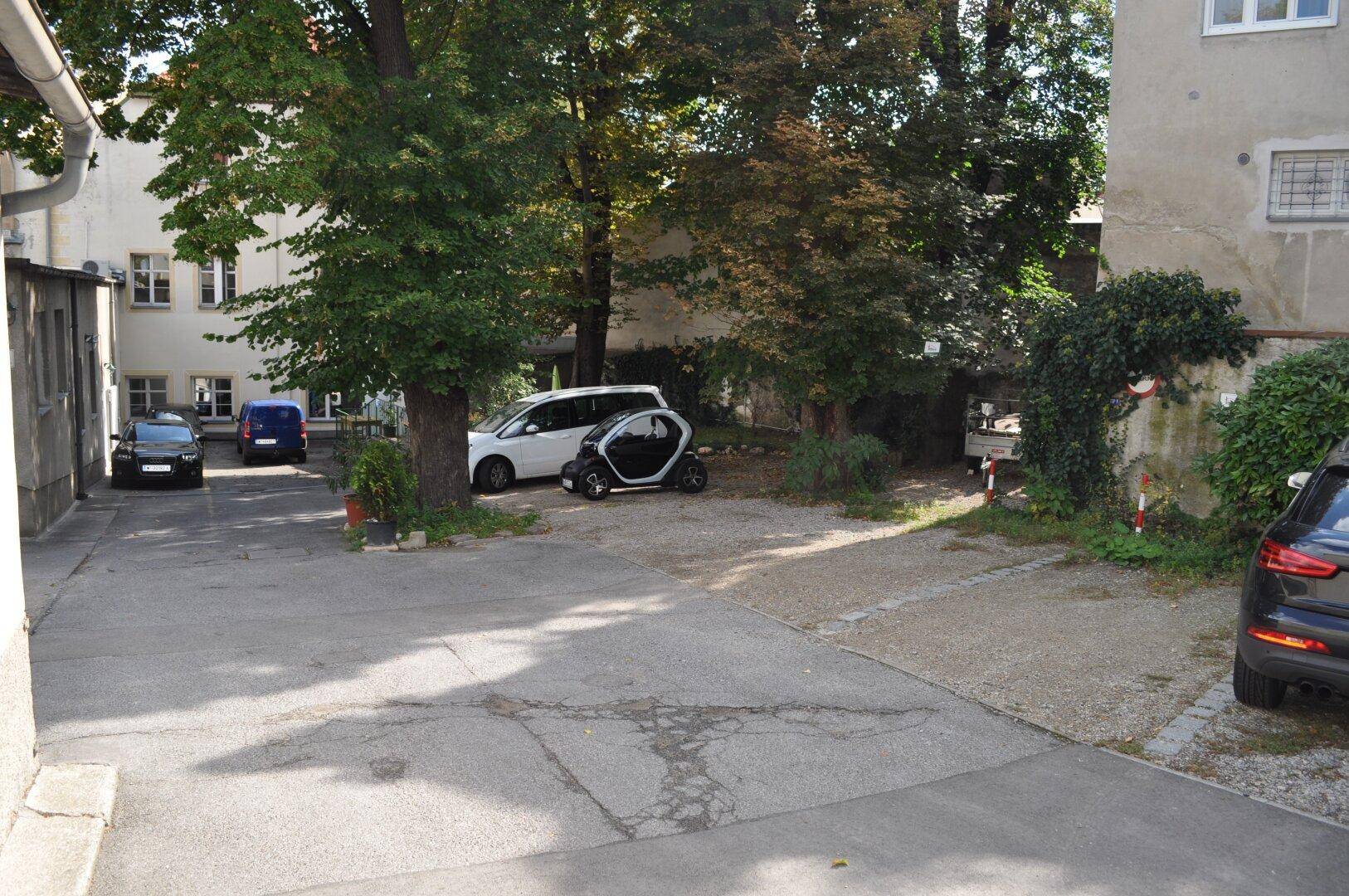Außenparkplatz mit mehreren Fahrzeugen und Bäumen, umgeben von Gebäuden in einem Hinterhofbereich.
