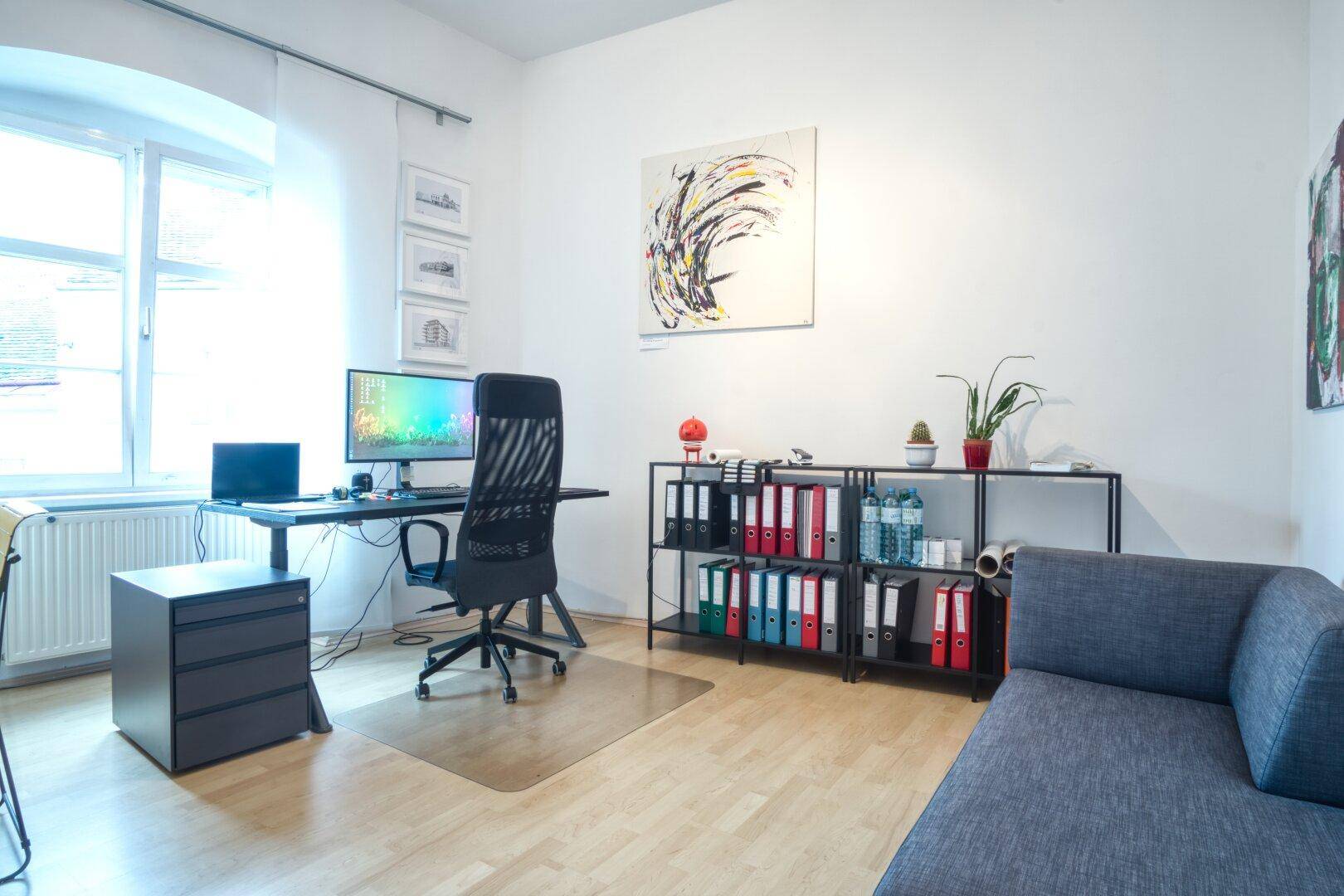 Helles Büro mit großem Fenster, Schreibtisch, Bürostuhl und einem Regal voller Aktenordner.