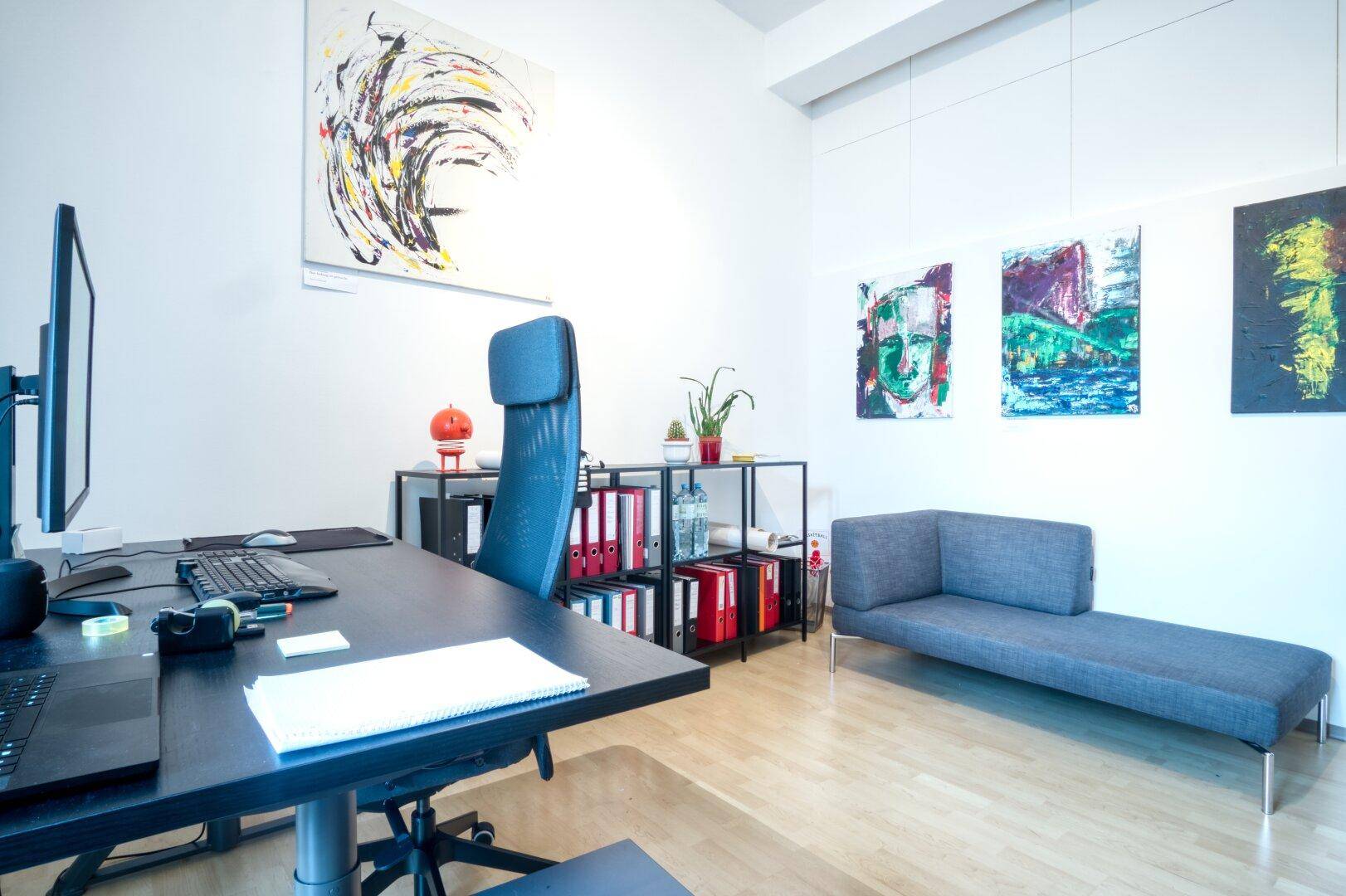 Modern eingerichtetes Büro mit Schreibtisch, ergonomischem Stuhl und einer grauen Chaiselongue, ergänzt durch Kunstwerke.